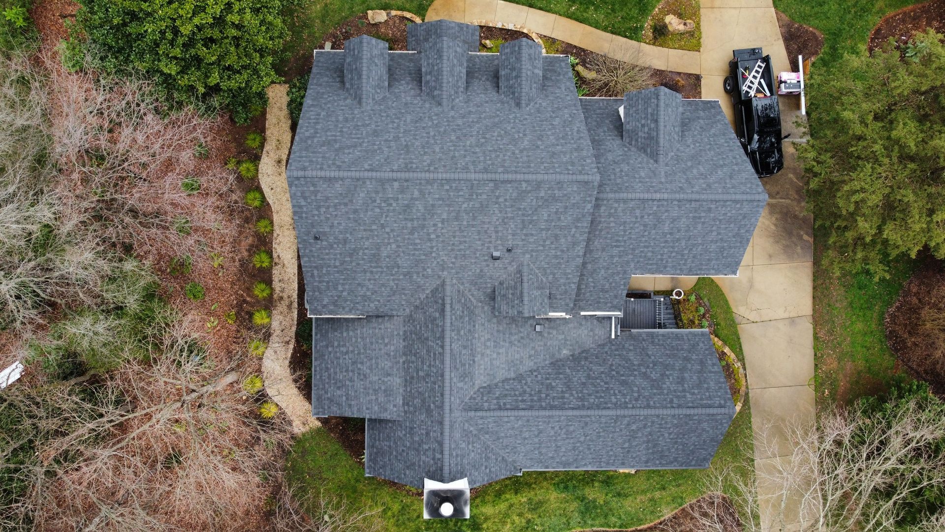 Aerial view of a gray-shingled house roof with surrounding trees and driveway.
