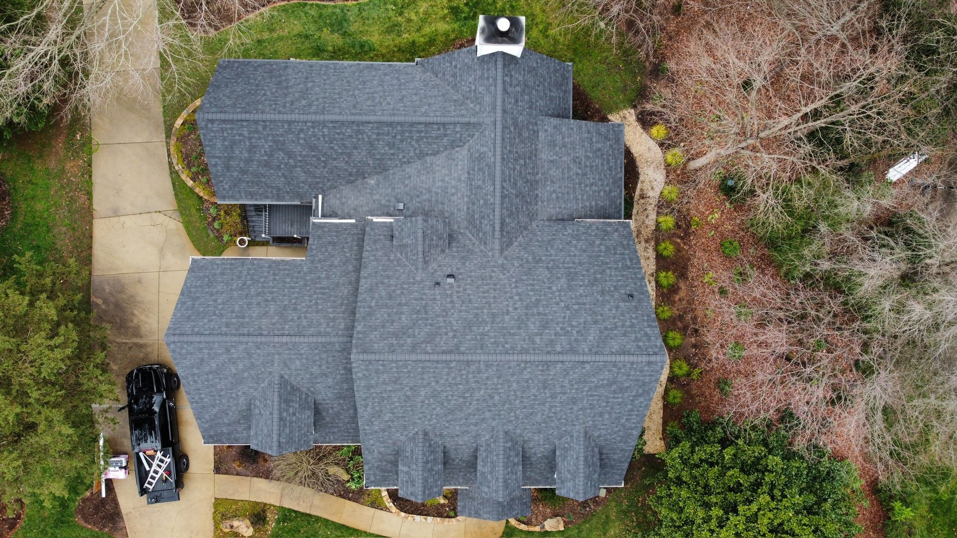 Aerial view of a gray-roofed house surrounded by trees and a driveway with two parked vehicles.