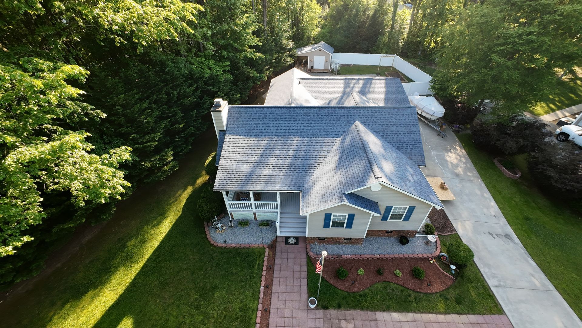 Aerial view of a small blue house with a red-brick walkway, driveway, and trees surrounding it