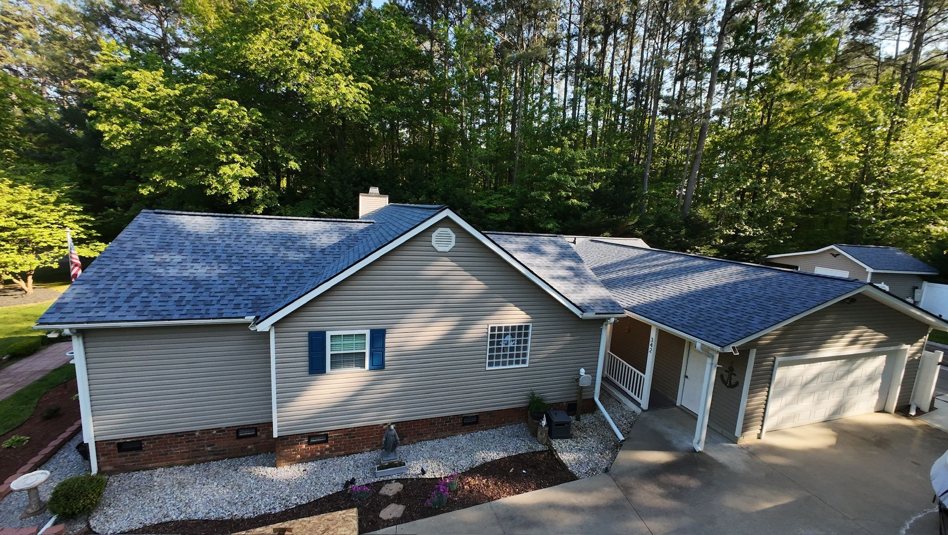 Aerial view of a gray house with blue roof, attached garage, and wooded yard in daylight