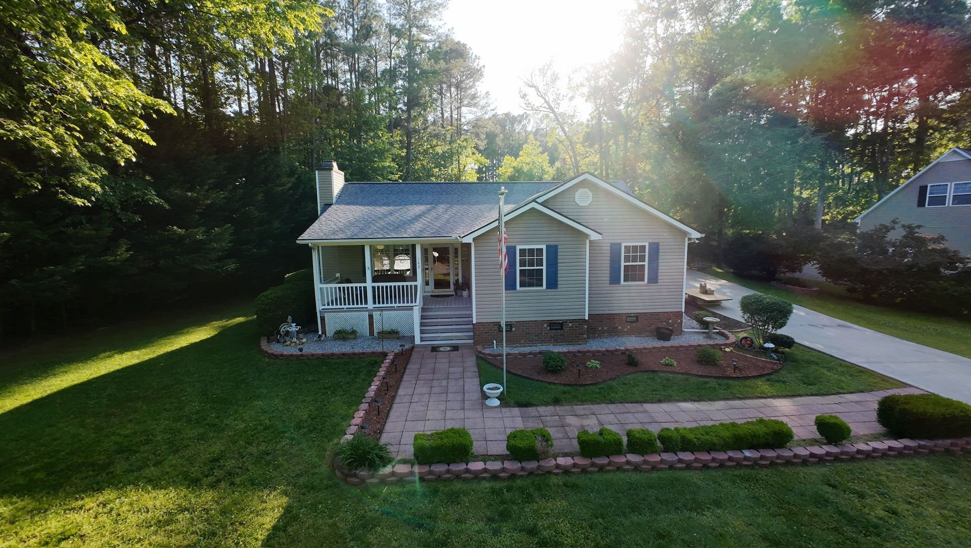 Small single-story house with a front porch, brick walkway, and sunlit green yard surrounded by trees.