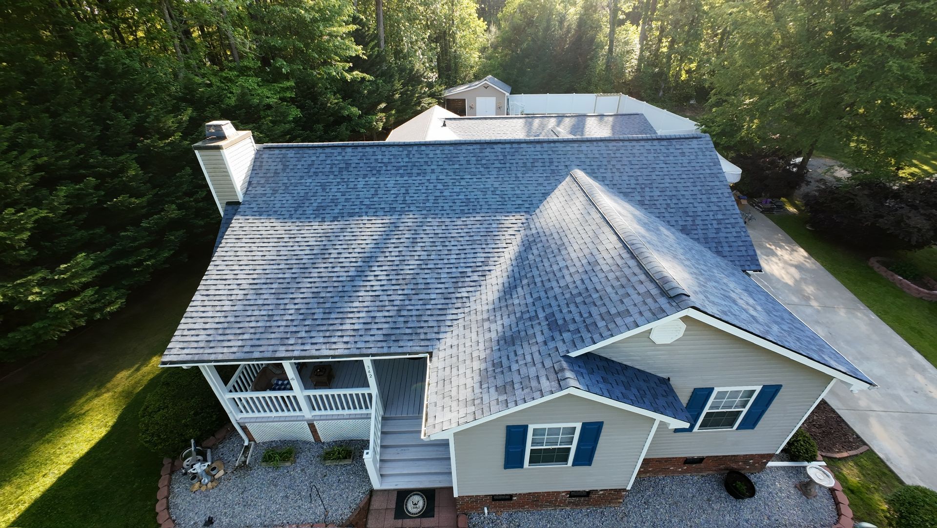 Aerial view of a beige house with a gray shingled roof, surrounded by trees and a gravel driveway.