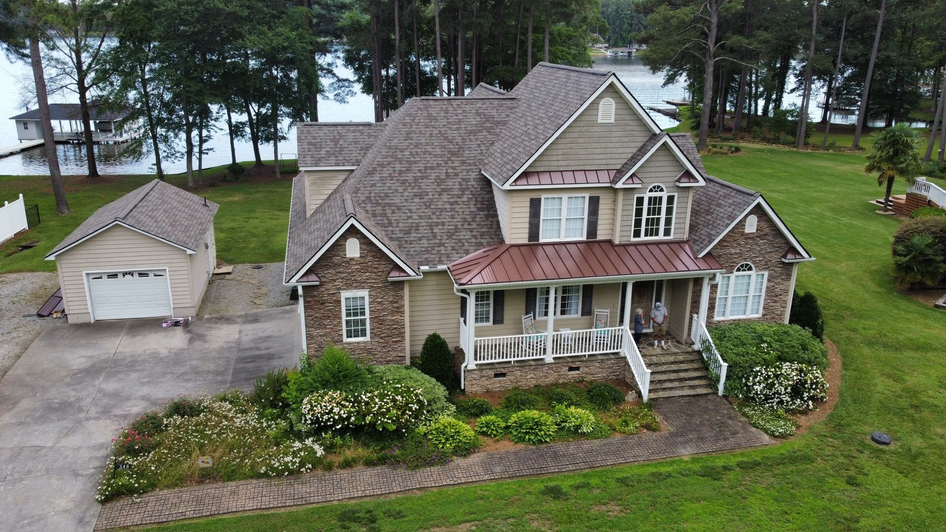 Aerial view of a stone house with a detached garage, front porch, and lakeside yard.