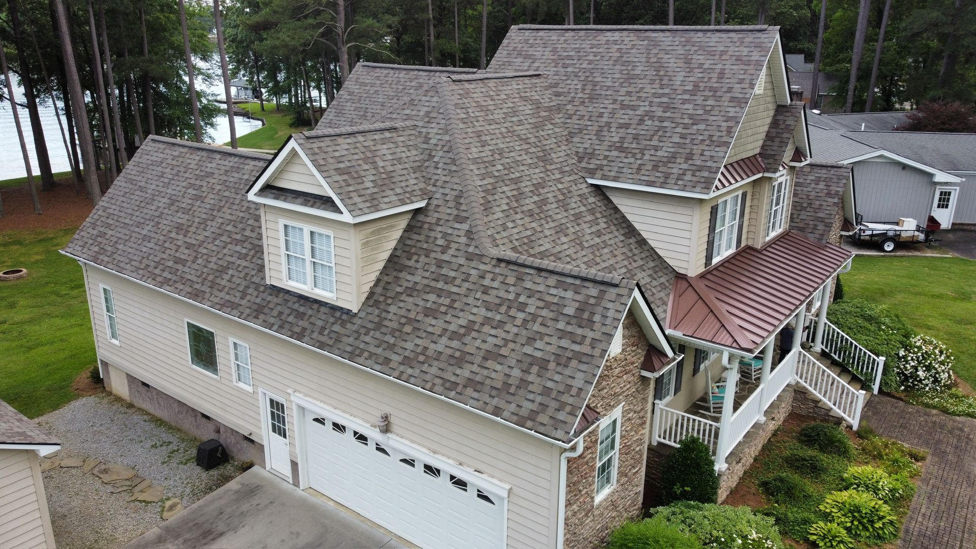 Aerial view of a large tan house with a gray roof, white garage, and red porch awning near trees