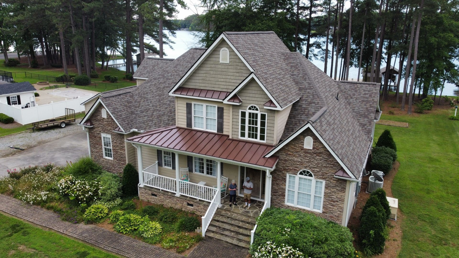Large brick house with a gray roof and front porch, surrounded by trees and a green lawn near water.