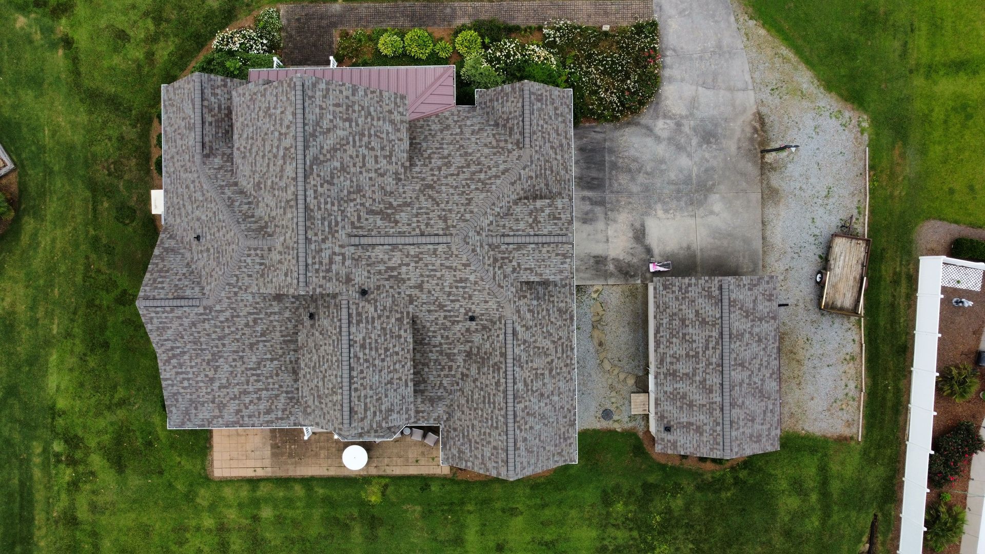 Aerial view of a house with a gray shingled roof, driveway, and surrounding green yard.