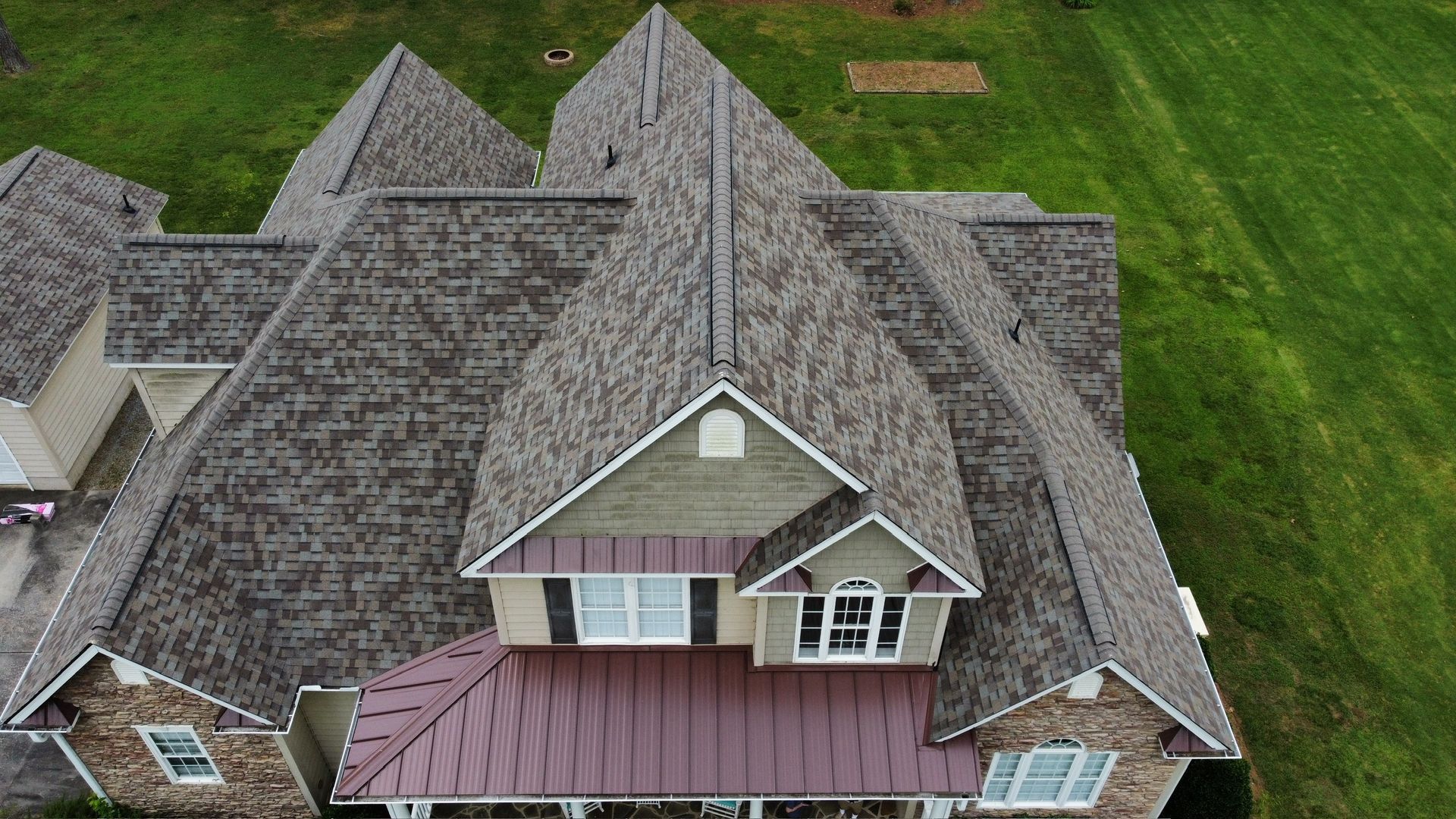 Aerial view of a large house with multiple gray shingled roofs and a front porch on a green lawn