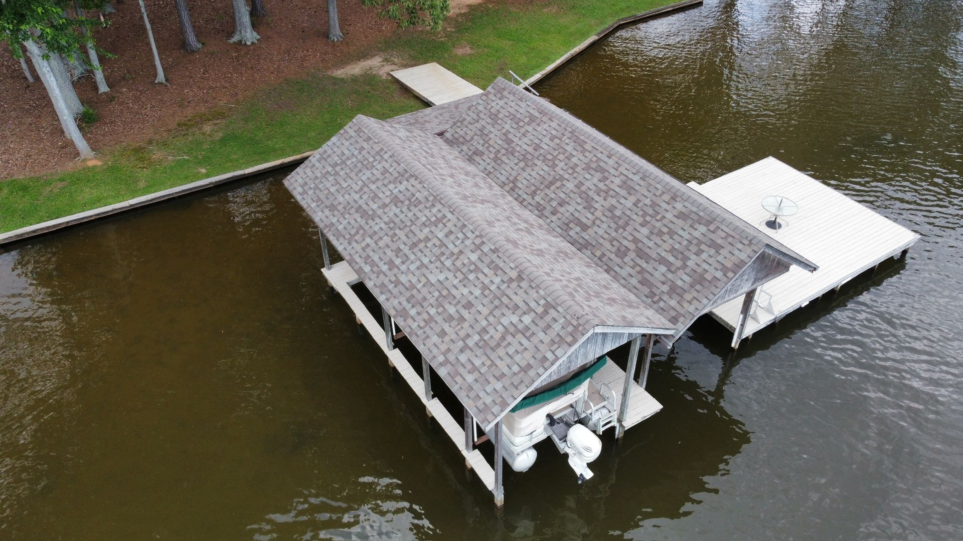Aerial view of a covered dock with a boat lift on a lake beside a grassy shoreline