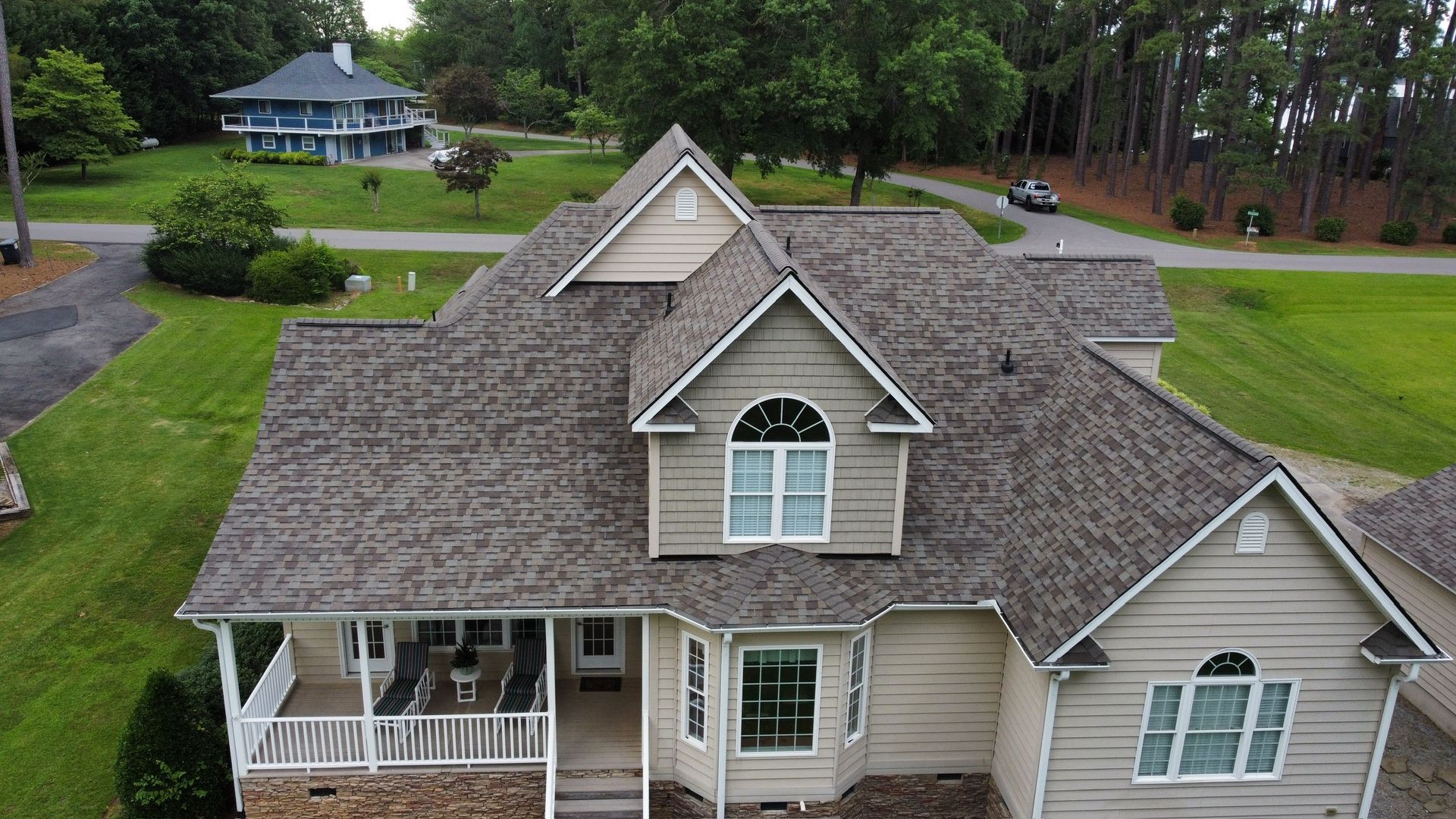 Aerial view of a beige suburban house with a gray shingle roof and front porch surrounded by green lawn.