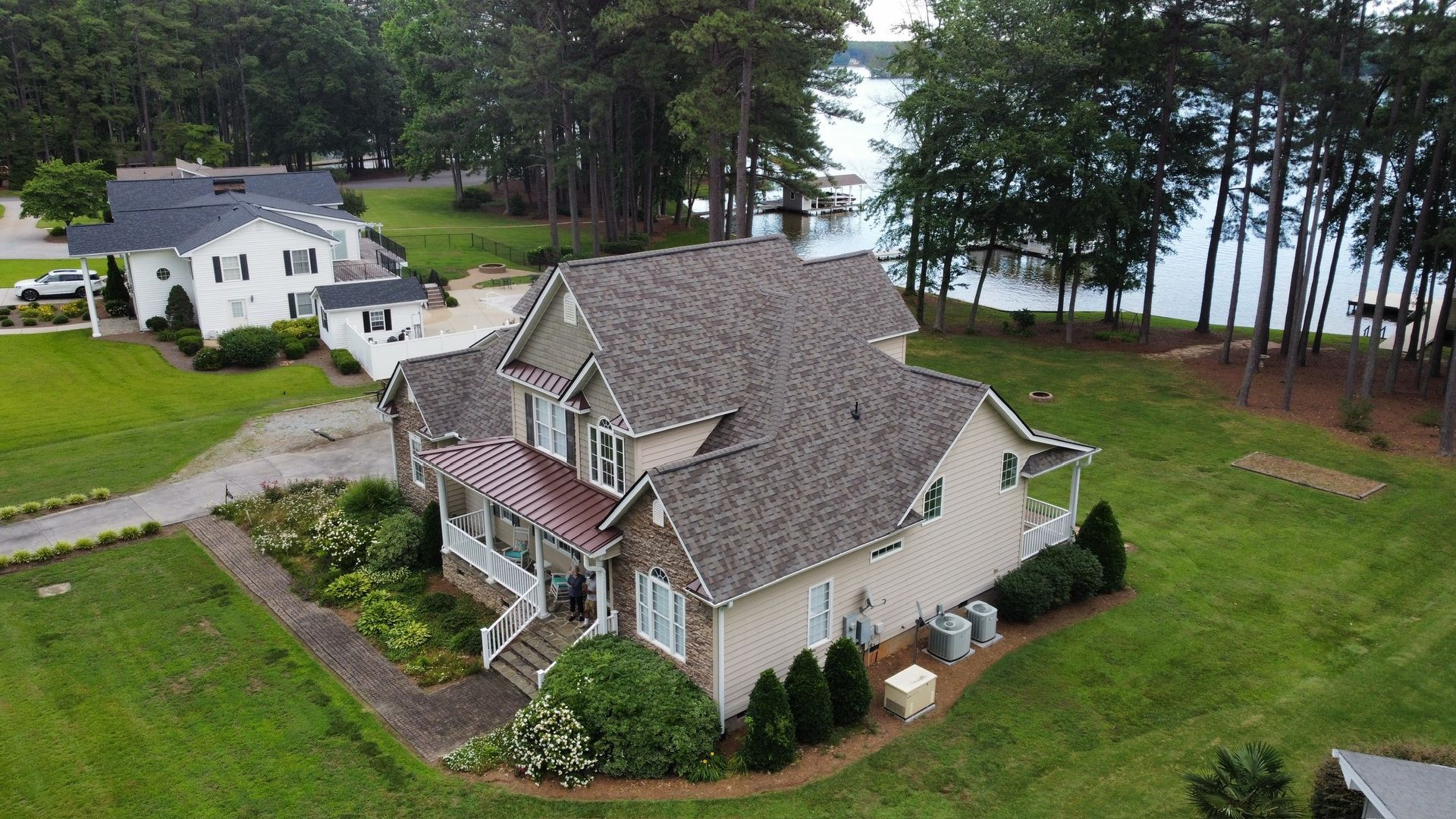 Aerial view of a large beige house with a gray roof near trees and water, with a smaller white house nearby