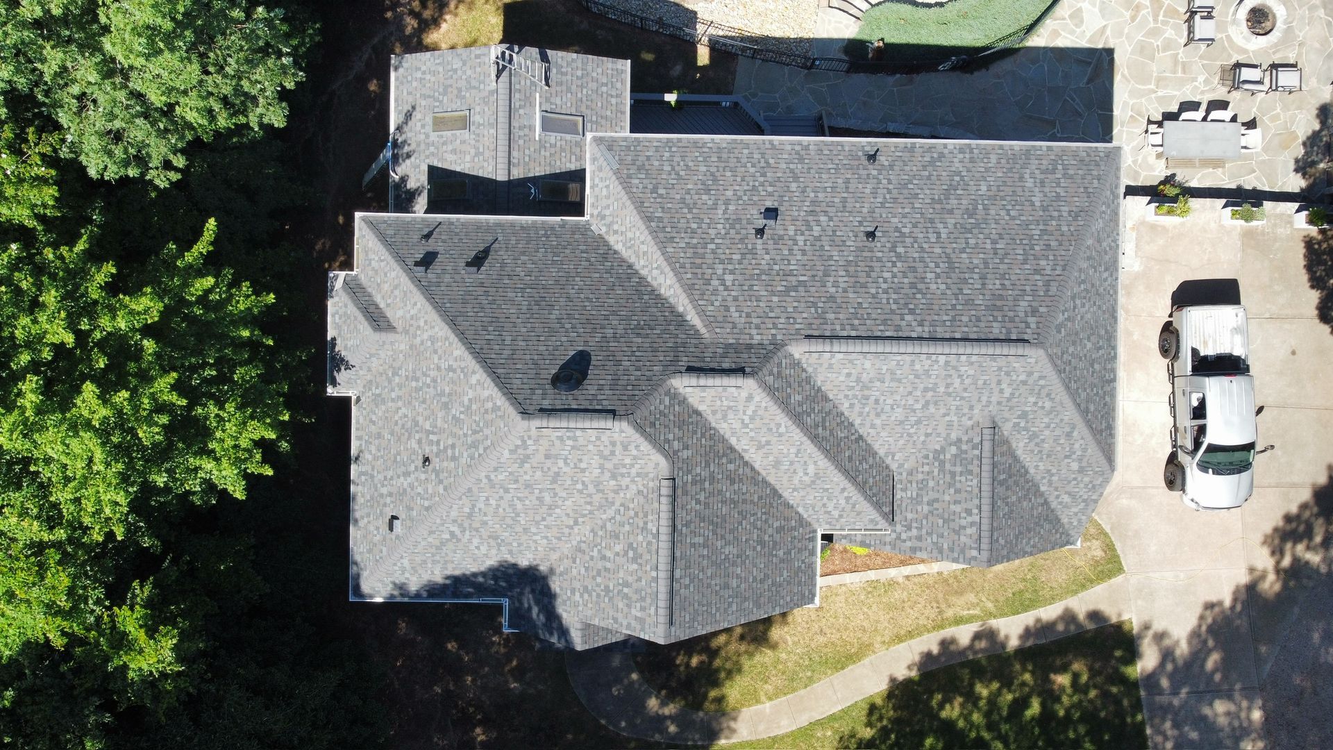 Aerial view of a gray-roofed house with a driveway, parked cars, and trees nearby.