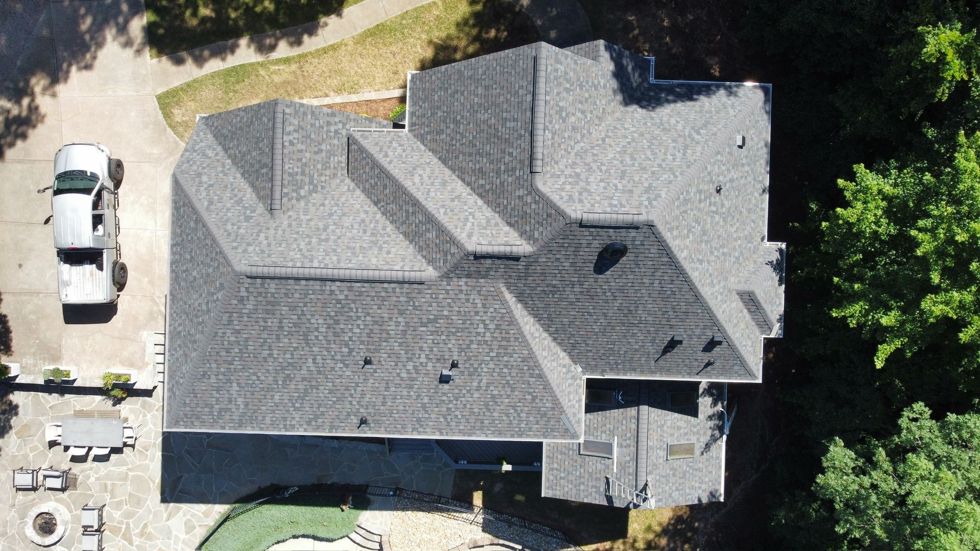 Aerial view of a gray-roofed house with surrounding trees and a driveway with a white vehicle.