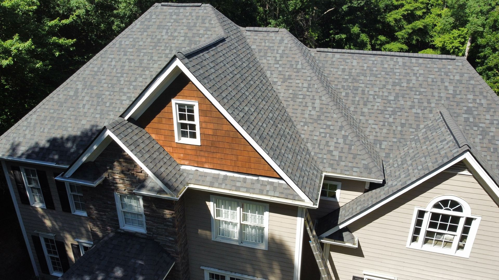 Aerial view of a house with a gray shingled roof and brick gable siding surrounded by trees.