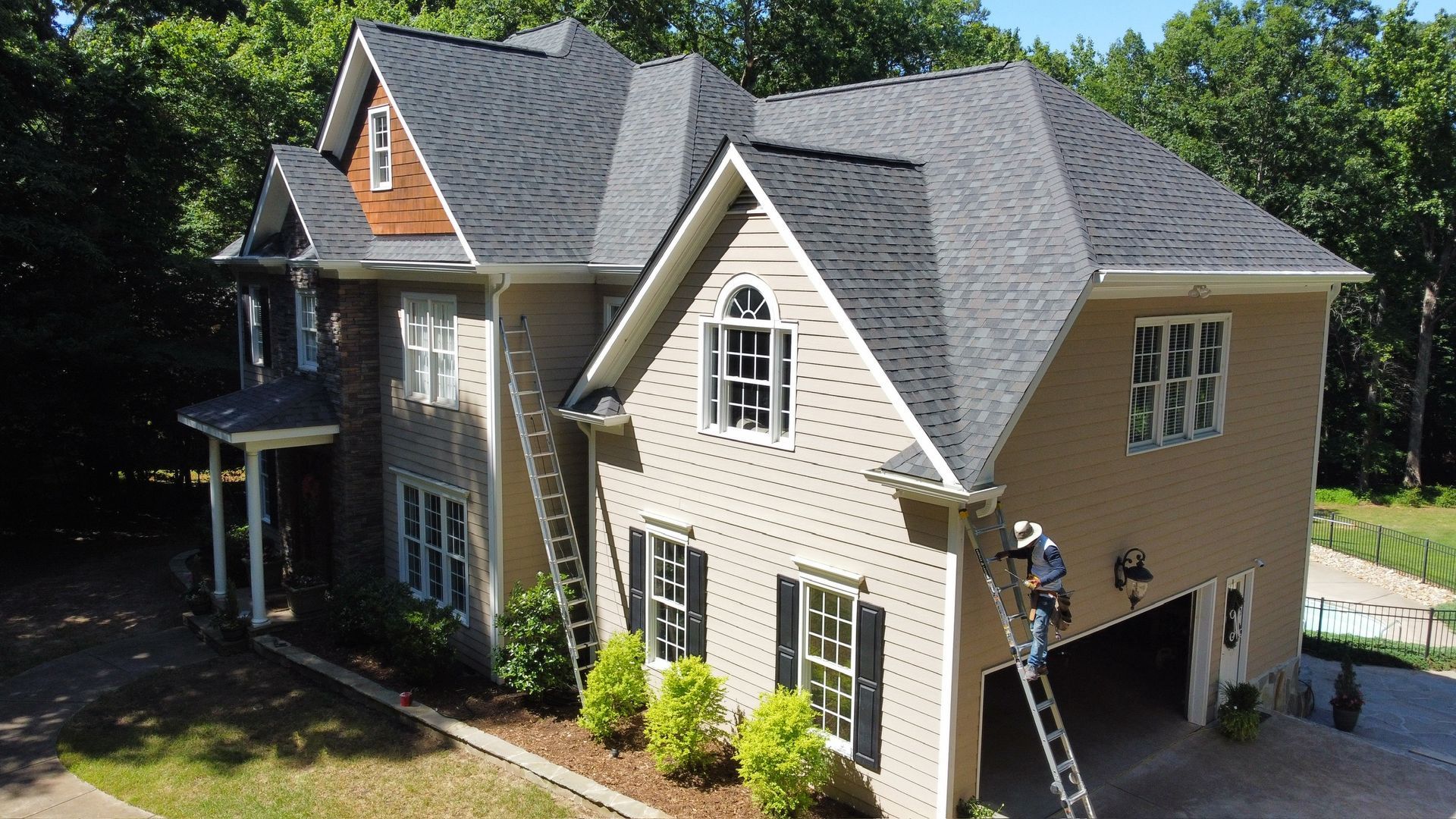 A two-story beige house with a gray roof and multiple windows, seen from above in a wooded neighborhood.