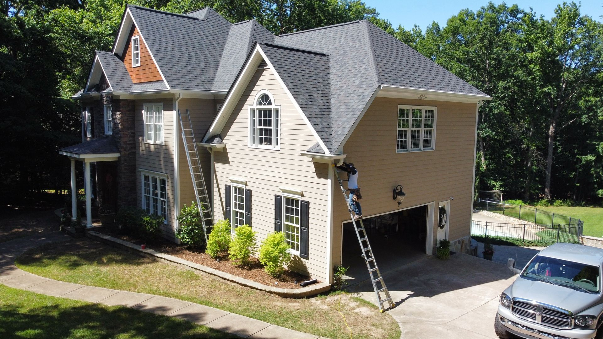 Two-story beige house with gray roof, front porch, driveway, and parked car in a leafy neighborhood