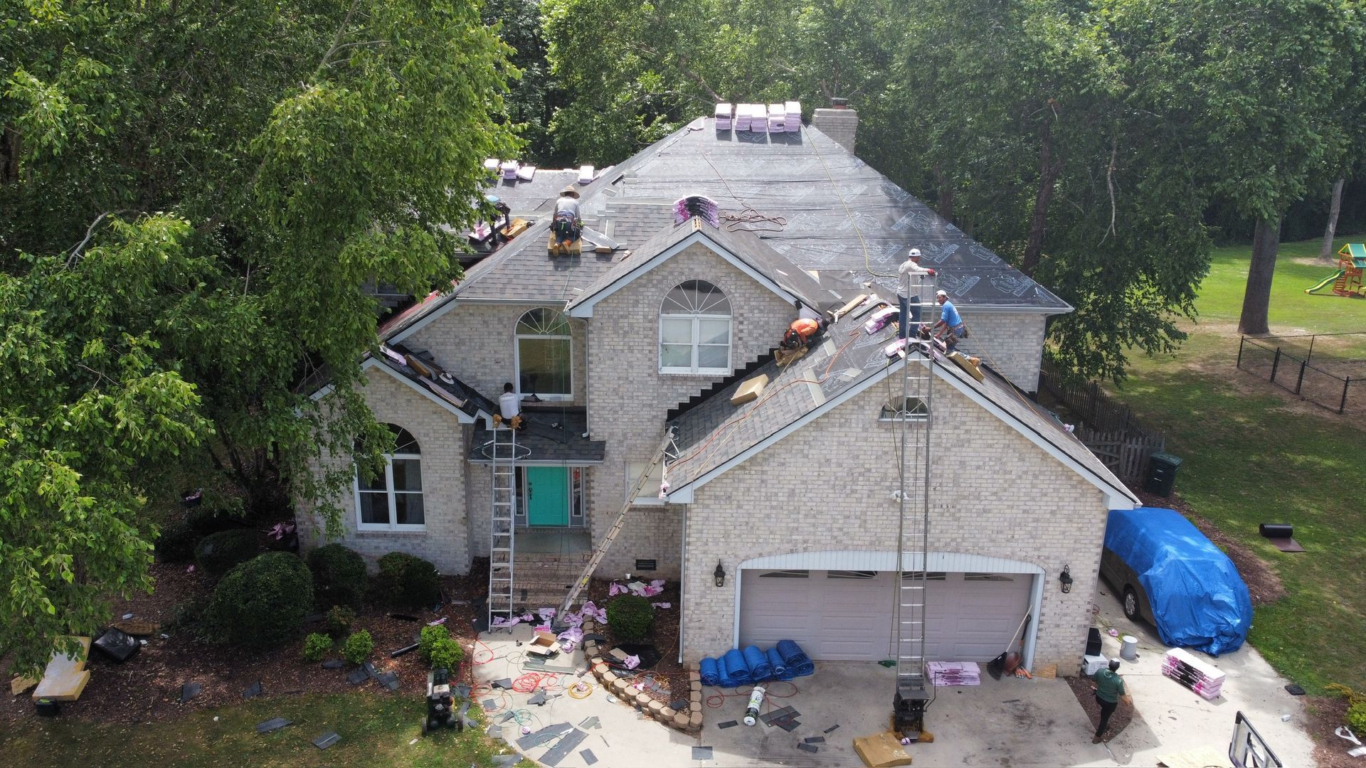 Aerial view of a house with roof repairs, tools and debris below, surrounded by trees.