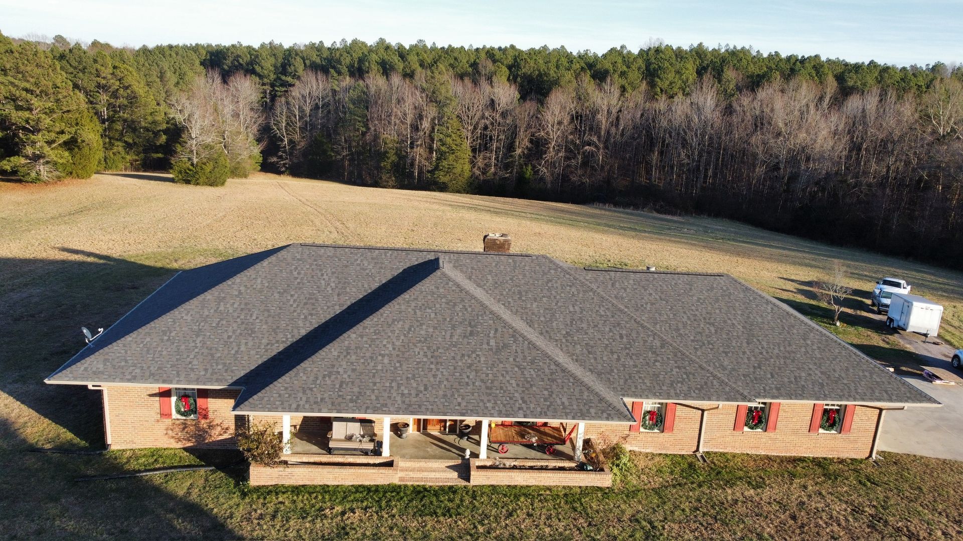 Aerial view of a single-story brick house with a dark roof, front porch, and wooded backyard.