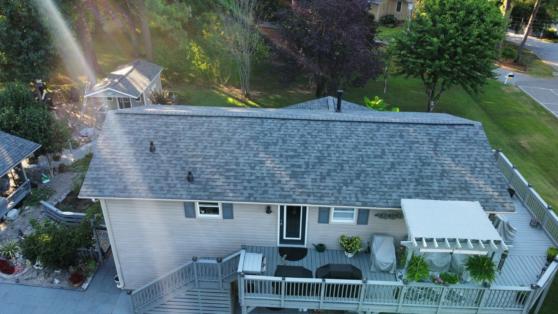 Aerial view of a light gray house with a front porch, white deck, and surrounding trees and yard.