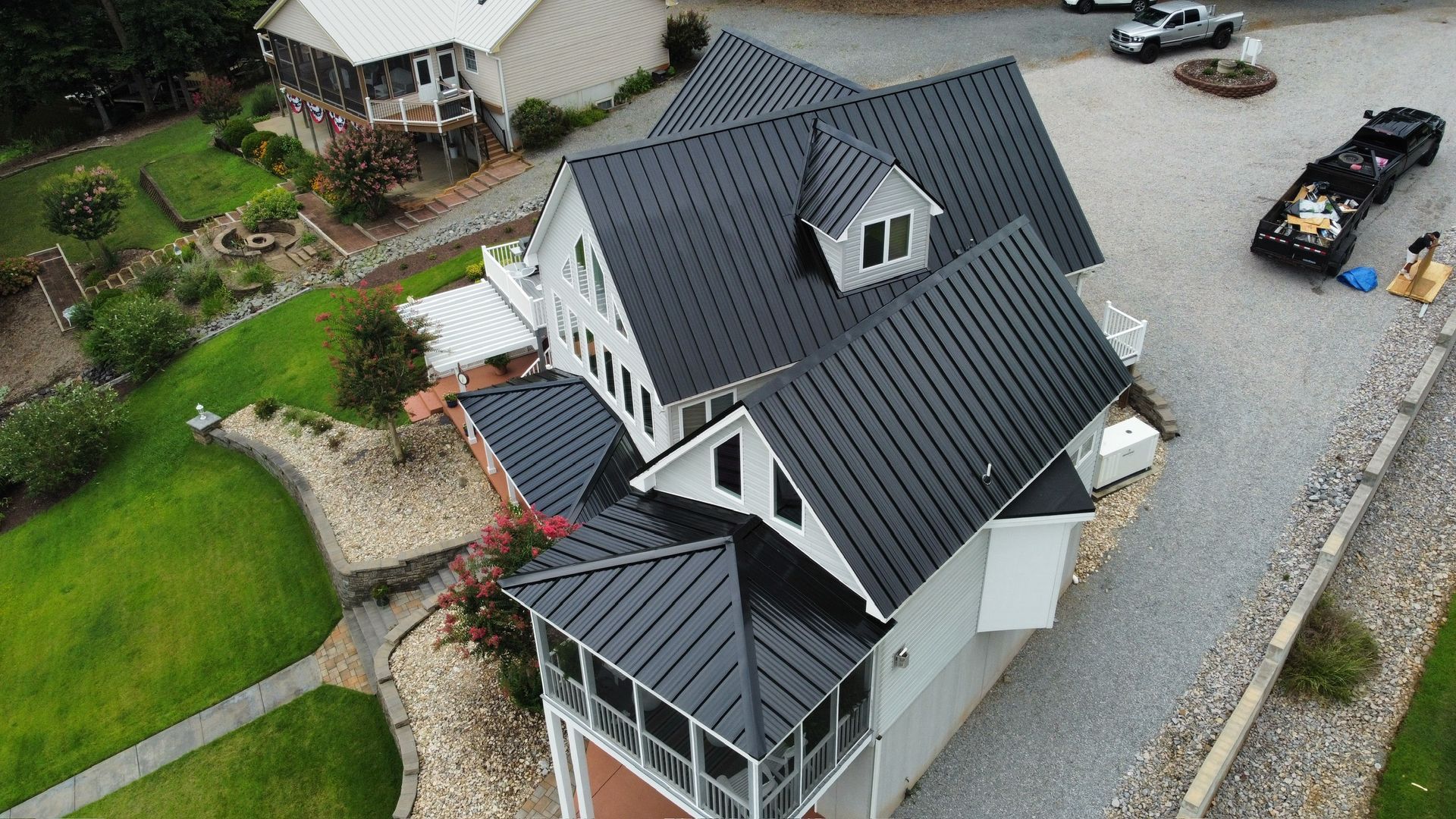 Aerial view of a dark gray house with a black roof, gravel driveway, and landscaped yard