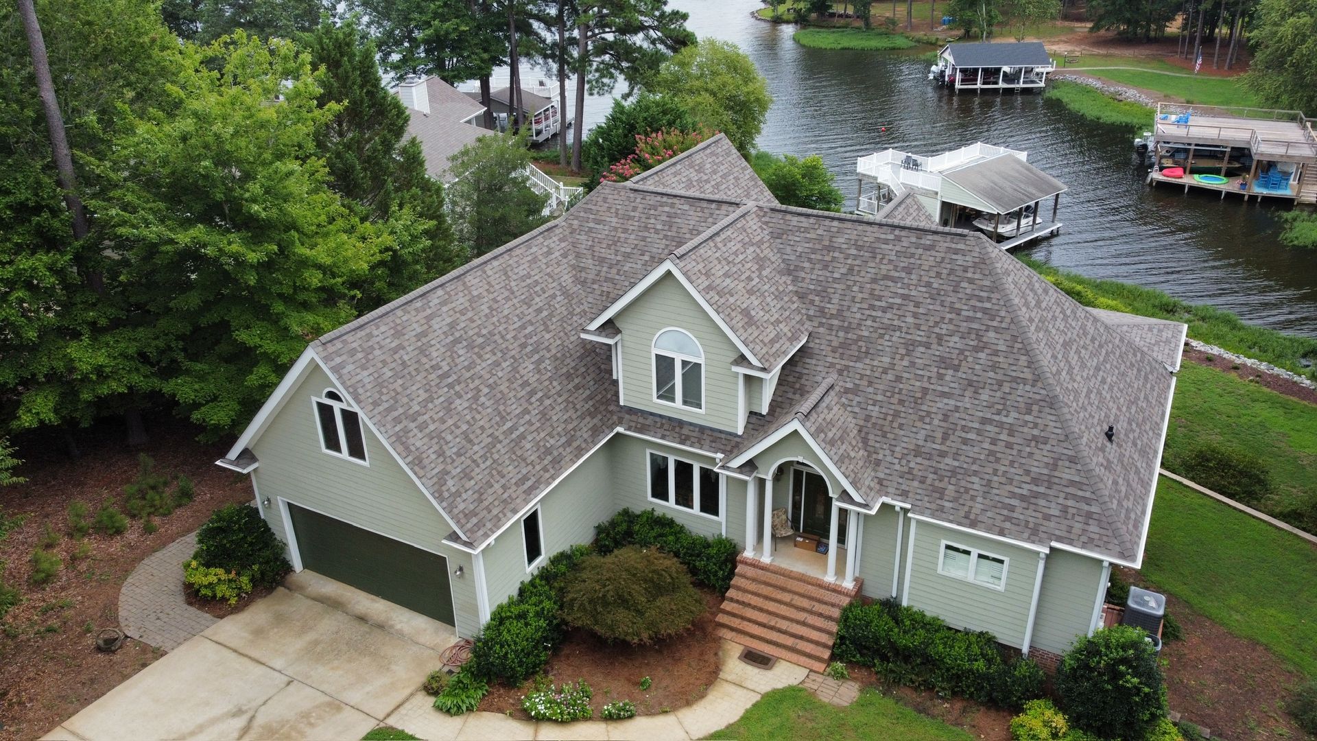 Aerial view of a gray house with a front porch beside a tree-lined lake with docks and boats.