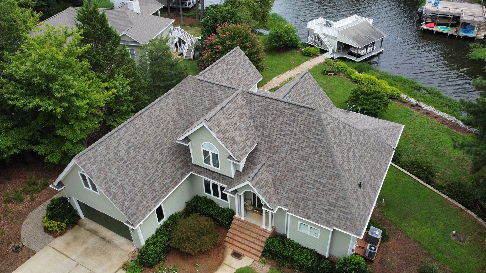 Aerial view of a gray house with a driveway, front steps, and a lush yard beside a waterfront dock.