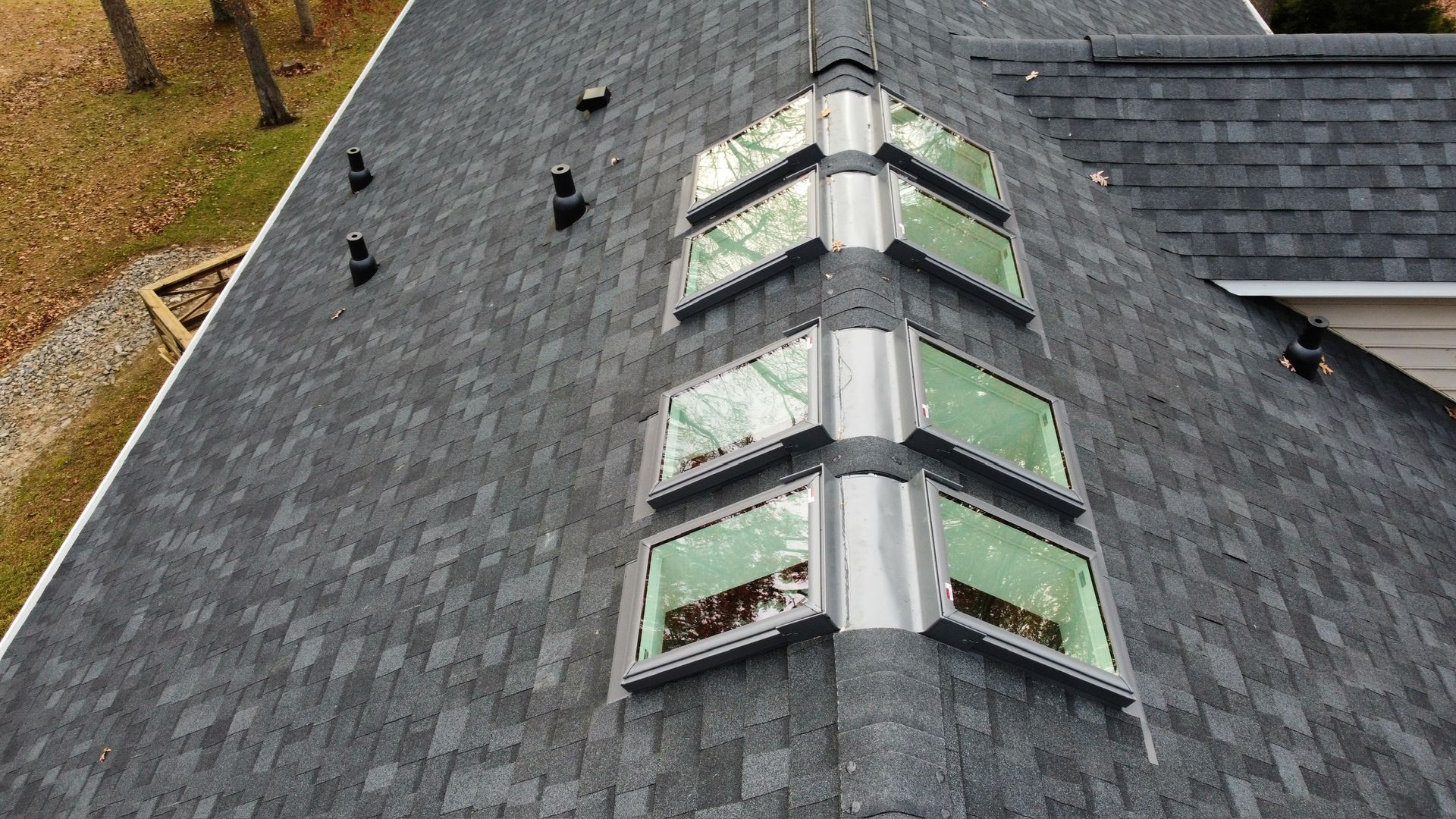 Aerial view of a dark shingled roof with a central skylight and several vents near a wooded area.