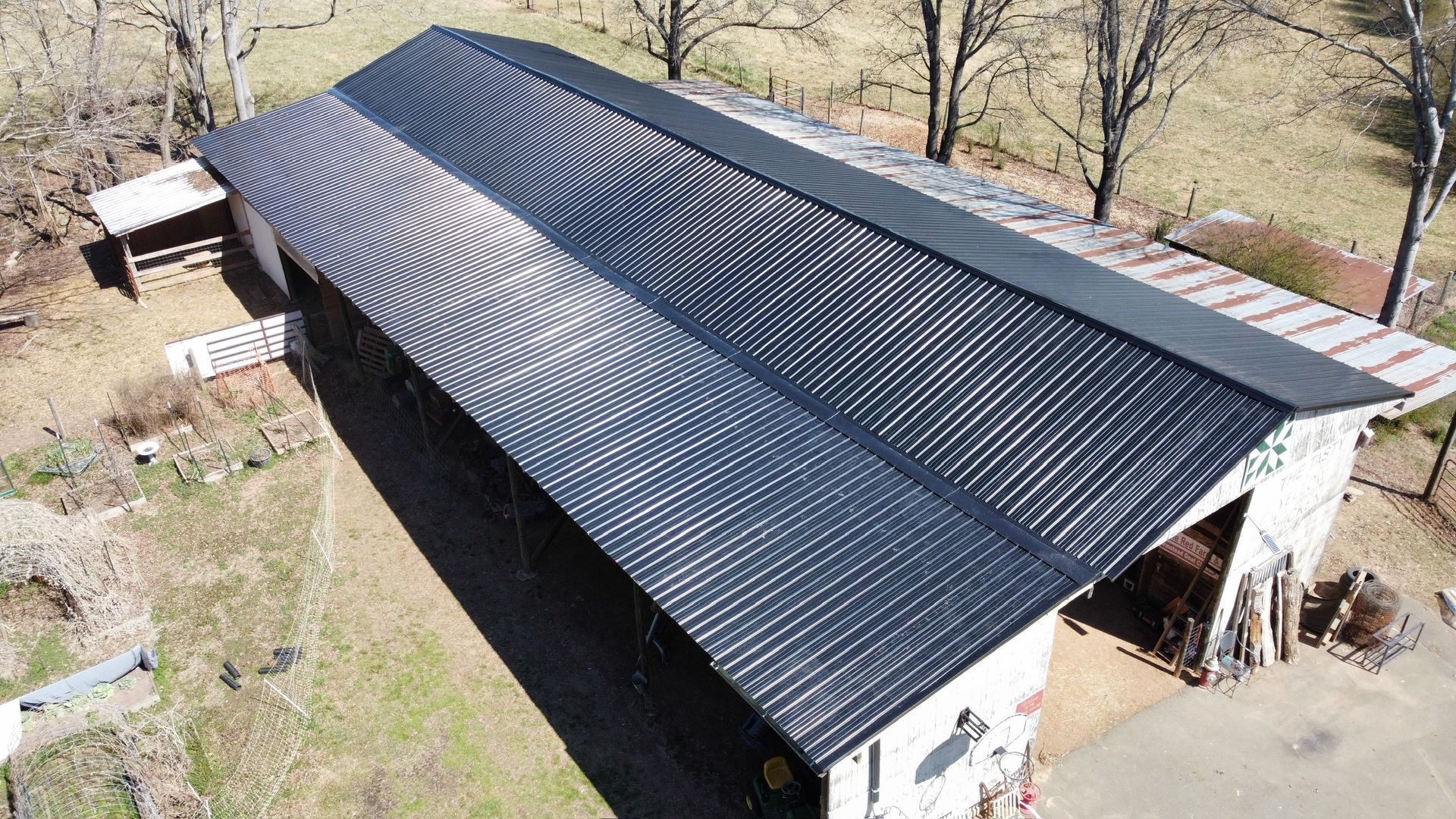 Aerial view of a large dark metal-roofed barn beside a grassy yard and leafless trees