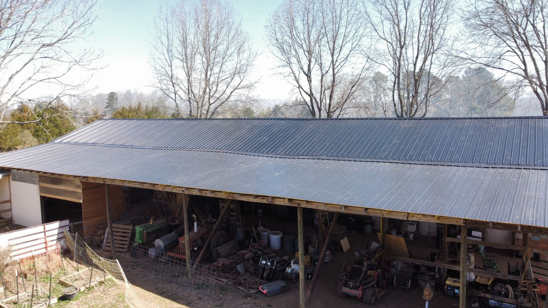 Long metal-roofed shed in a rural yard with leafless trees and stored items underneath