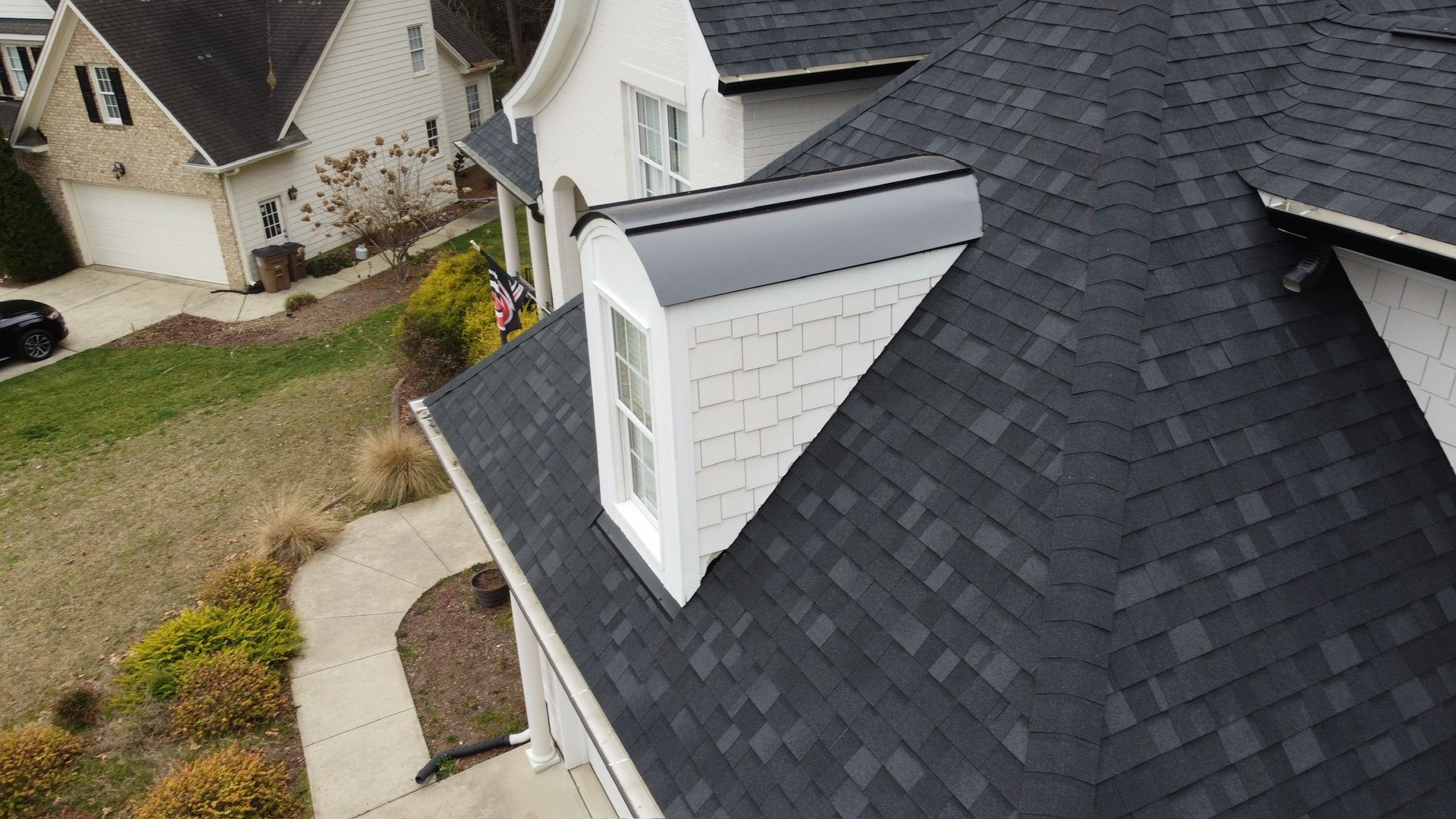 Aerial view of a house roof with a dormer and dark shingles beside a curved walkway and neighboring homes