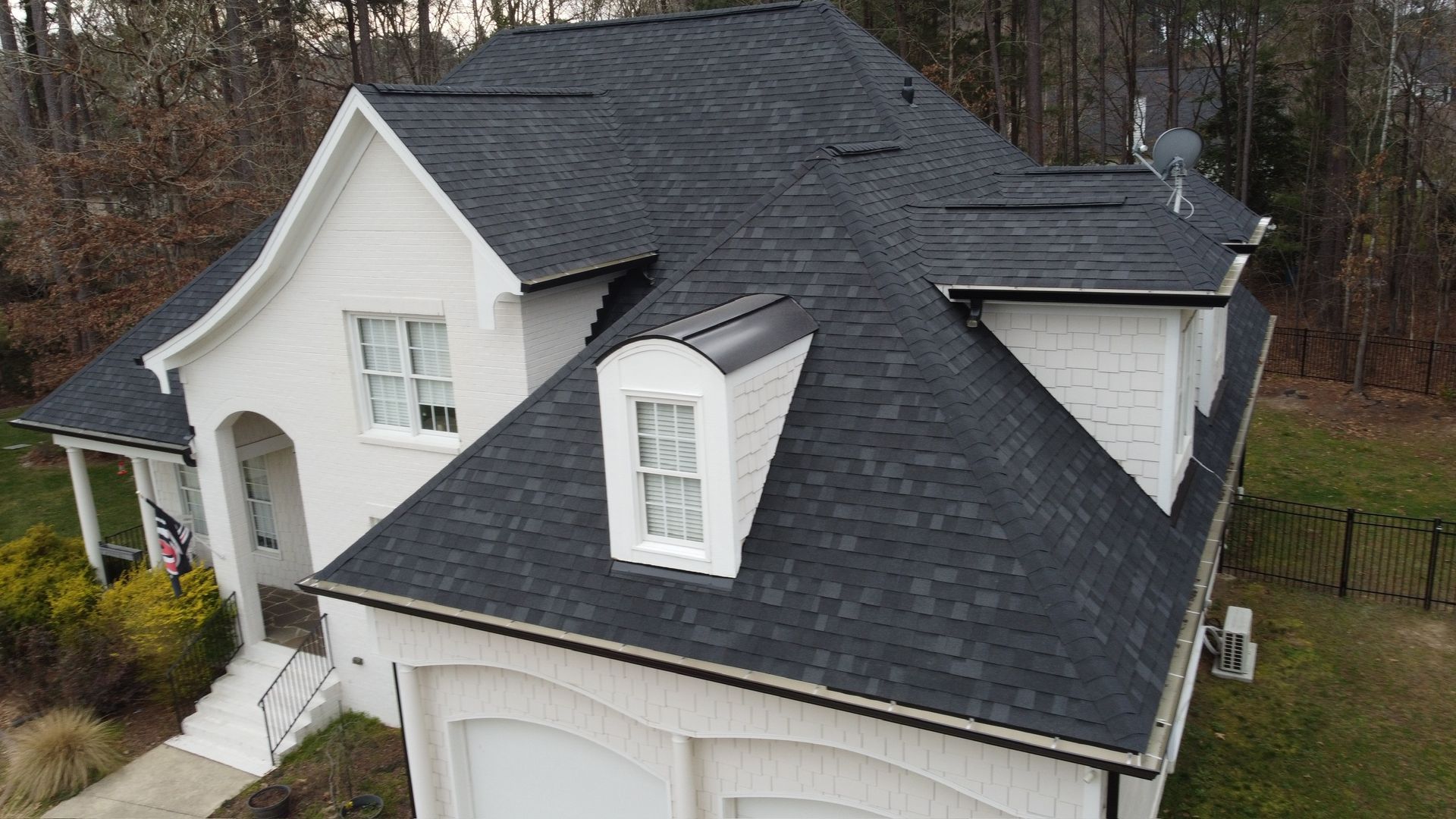 White house with dark gray roof and dormer windows, viewed from above in a wooded suburban setting