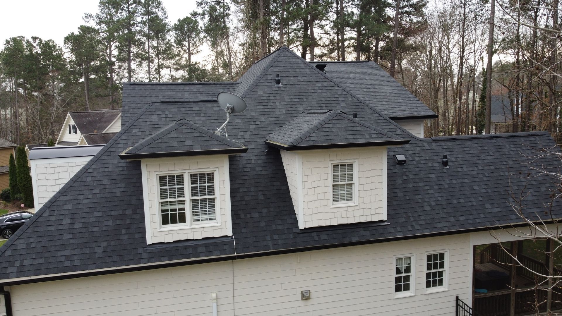 Dark gray shingled house roof with dormer windows and white siding, viewed from above with trees behind