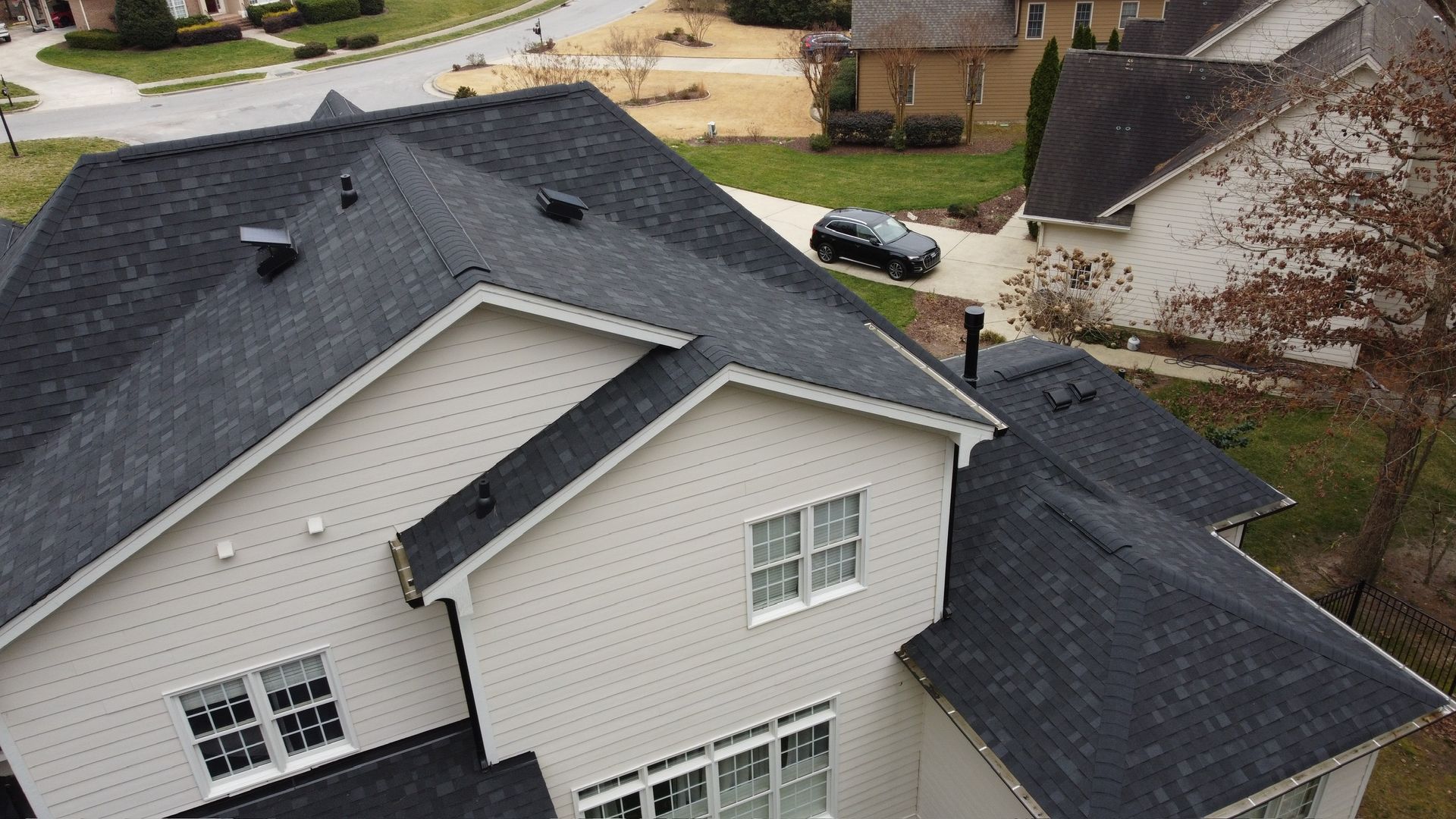 Aerial view of a suburban house with dark gray roof and light siding, near a driveway and street.