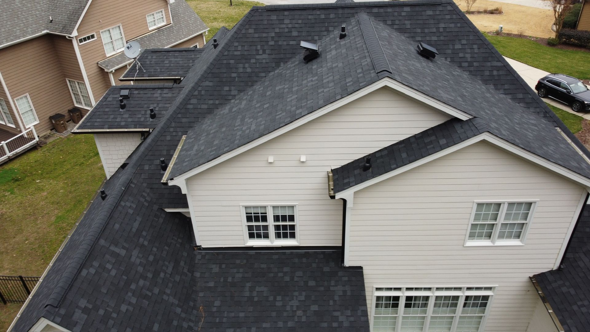Aerial view of a dark shingled house roof with light siding and dormer windows in a suburban neighborhood