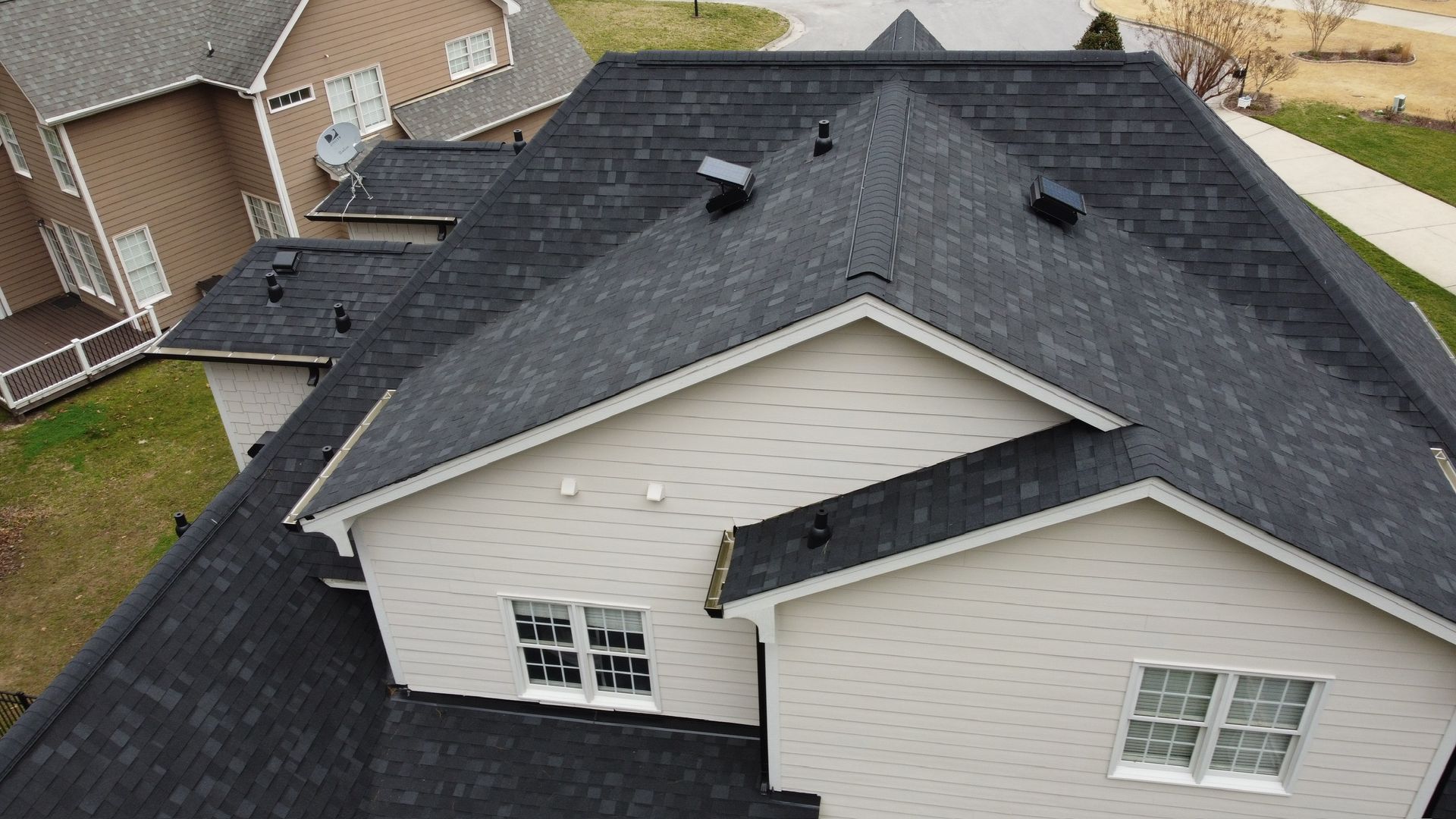 Aerial view of a dark shingle roof on a beige suburban house with multiple gables and dormer windows.