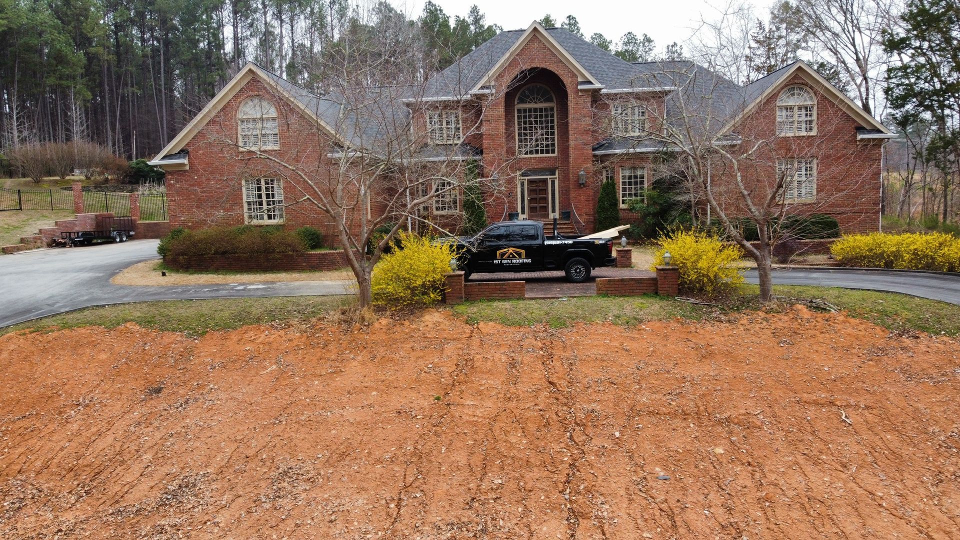 Large brick house with a black car in the driveway, surrounded by wet lawn and trees on a cloudy day