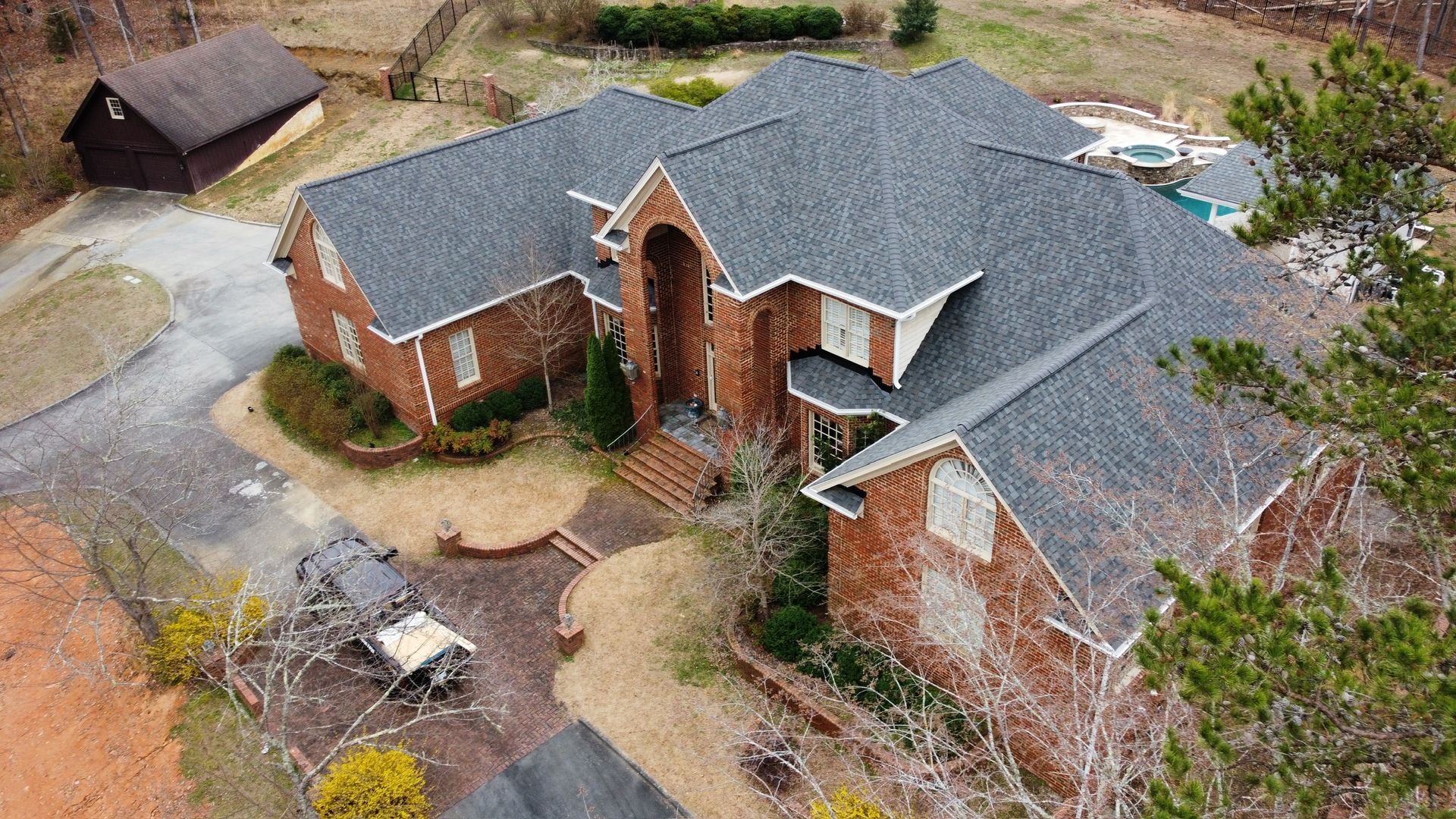 Aerial view of a large brick suburban house with a gray roof and curved driveway, surrounded by trees.