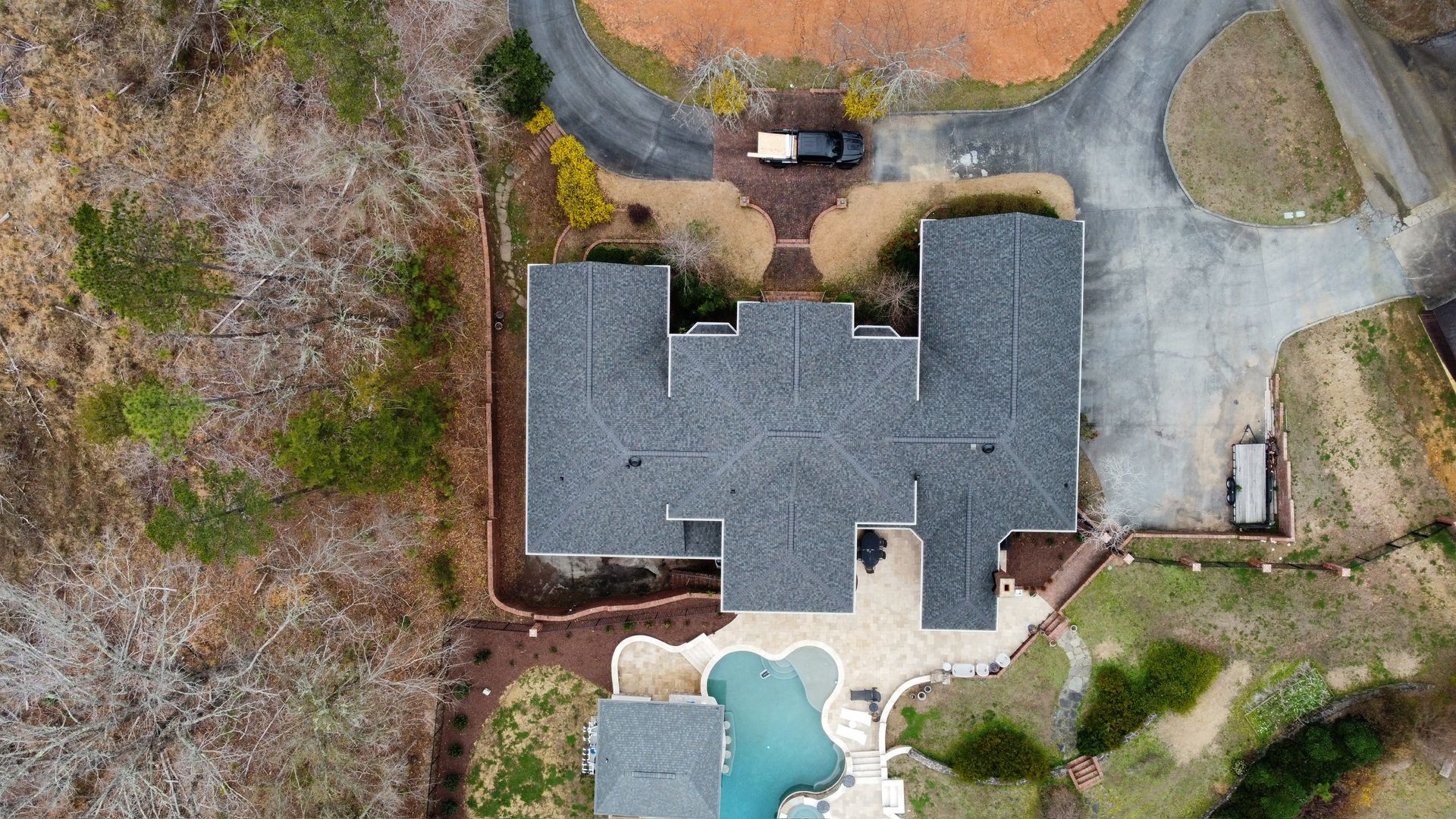 Aerial view of a house with a dark gray roof, driveway, and backyard pool surrounded by trees.