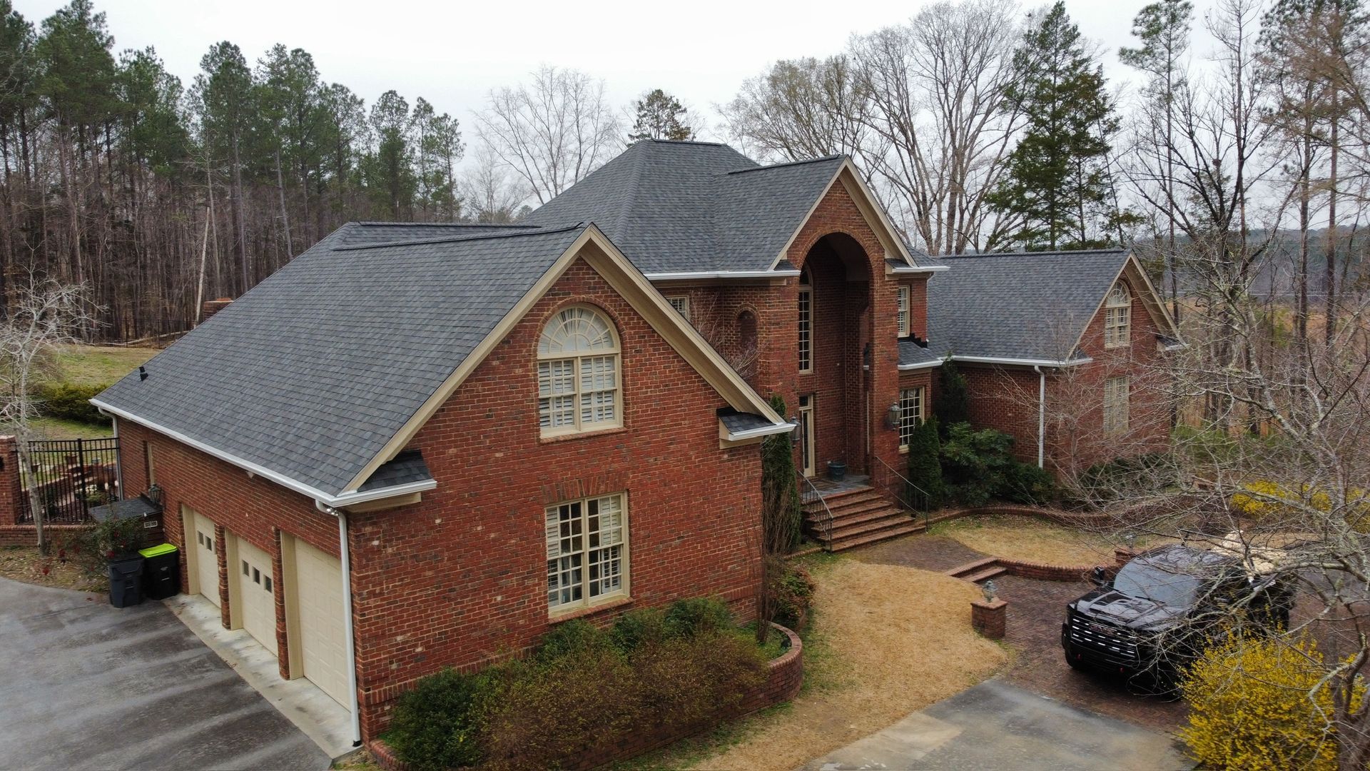 Brick suburban house with dark roof, attached garage, and wet driveway on a wooded lot