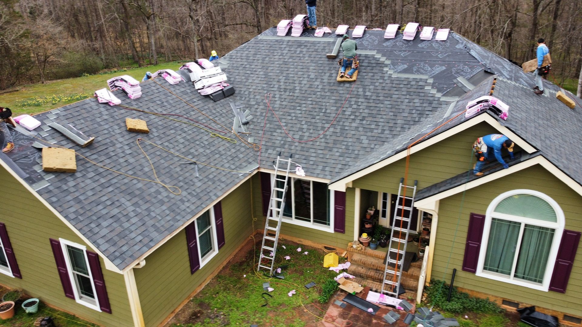 Roofers installing shingles on a gray roof of a house, with ladders and materials spread across the roof.