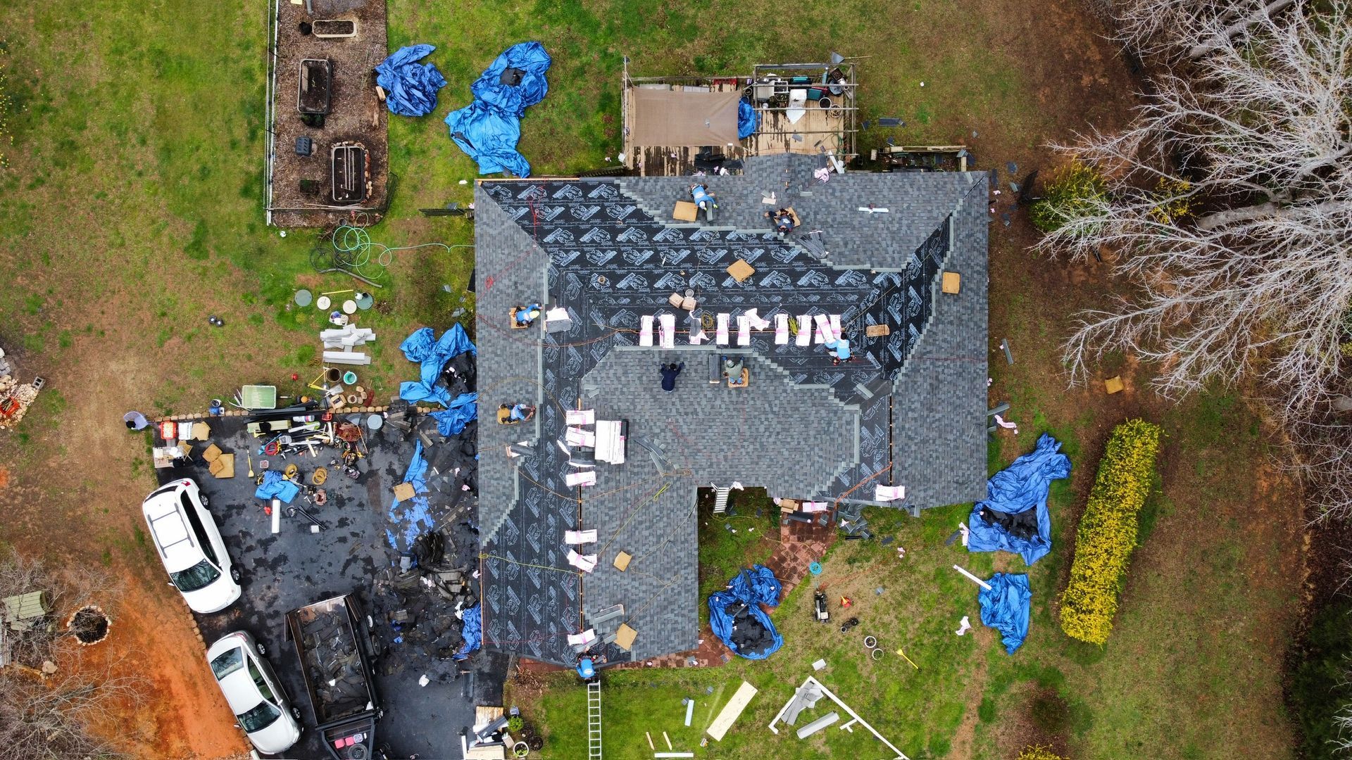 Aerial view of a house roof with blue tarps, debris, and a white truck in a muddy yard.