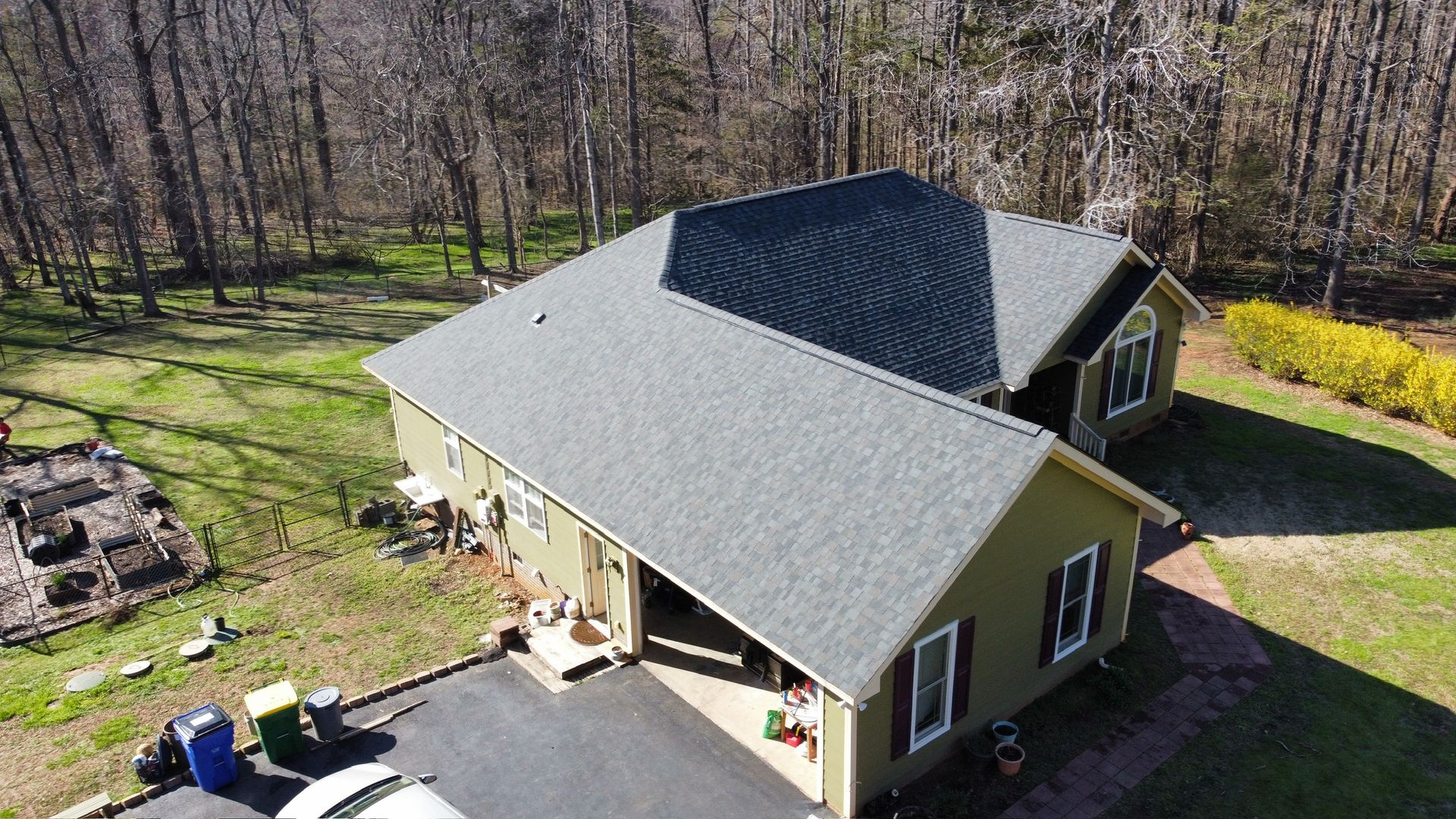 Aerial view of a beige house with a gray roof beside a driveway and wooded yard