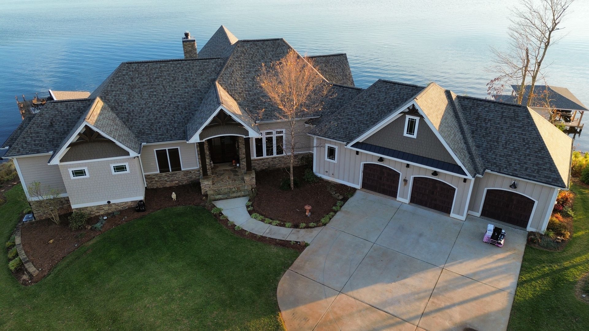 Aerial view of a large beige suburban house with a dark roof, curved driveway, and lawn beside water