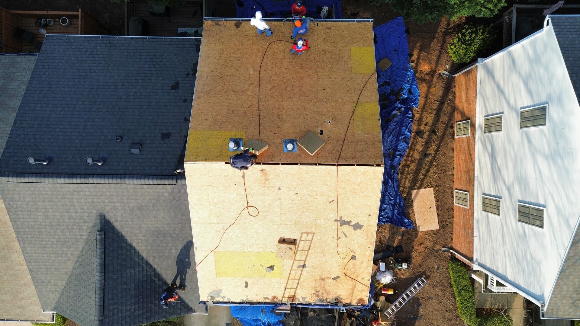 Aerial view of workers on a roof with two different shingle sections, beside neighboring houses.