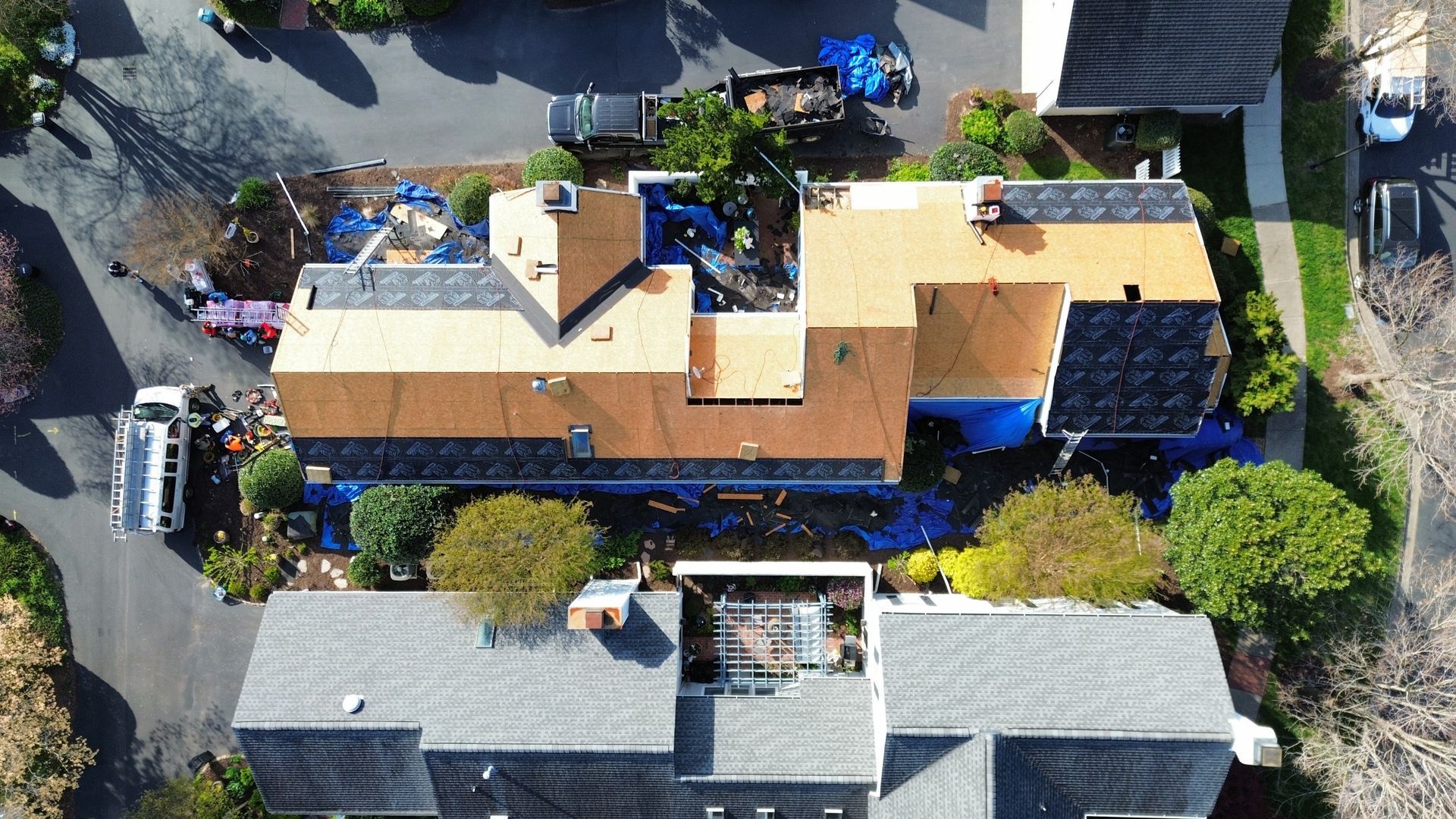 Aerial view of a house with tan roof, surrounded by trees, parked vehicles, and neighboring homes
