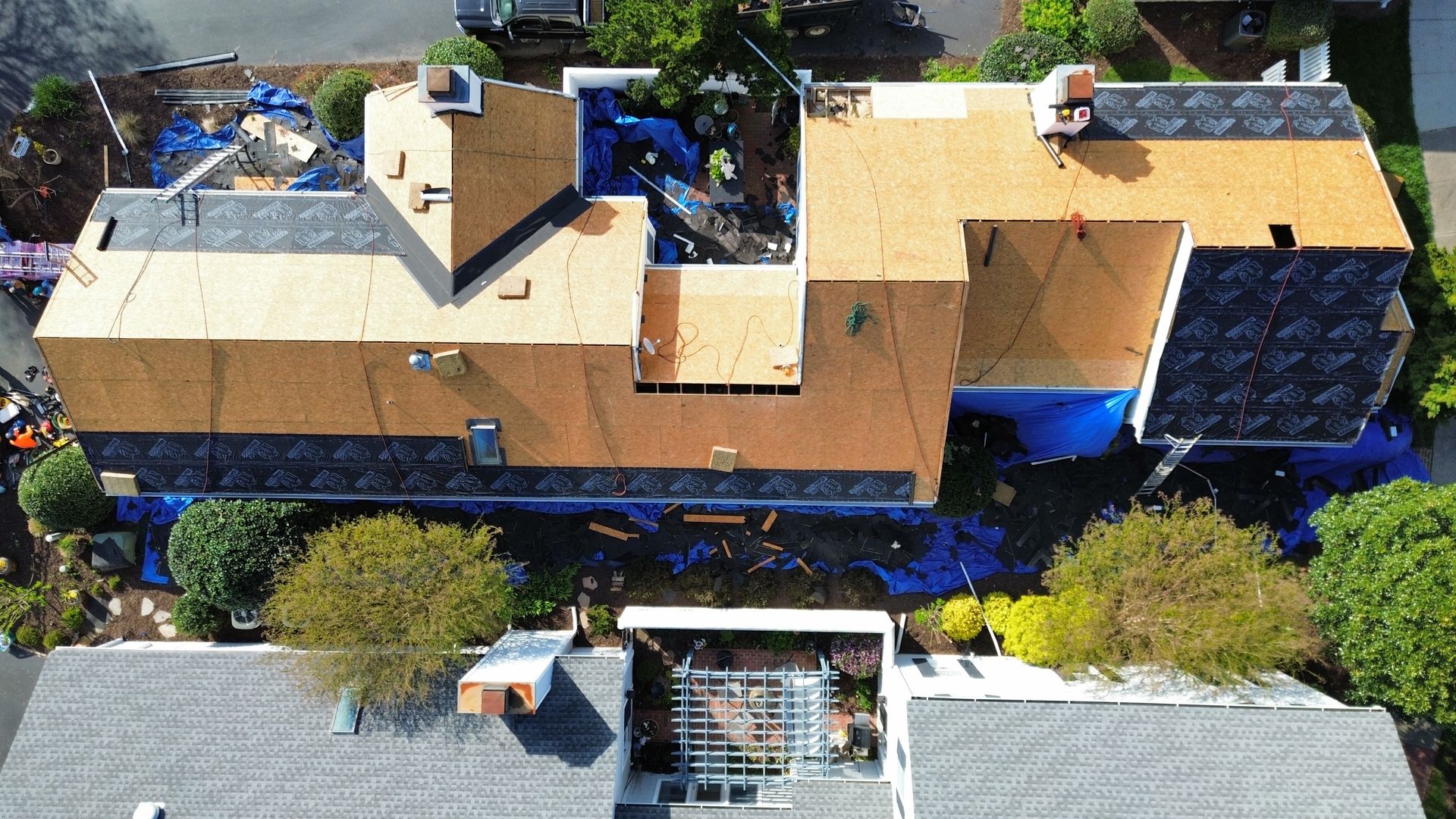 Aerial view of a large tan-roofed house with surrounding trees and neighboring rooftops.