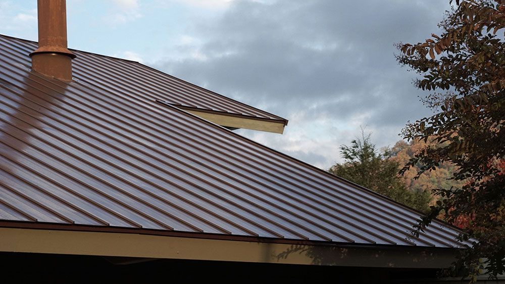Brown metal roof with a chimney under a cloudy sky, with trees in the background
