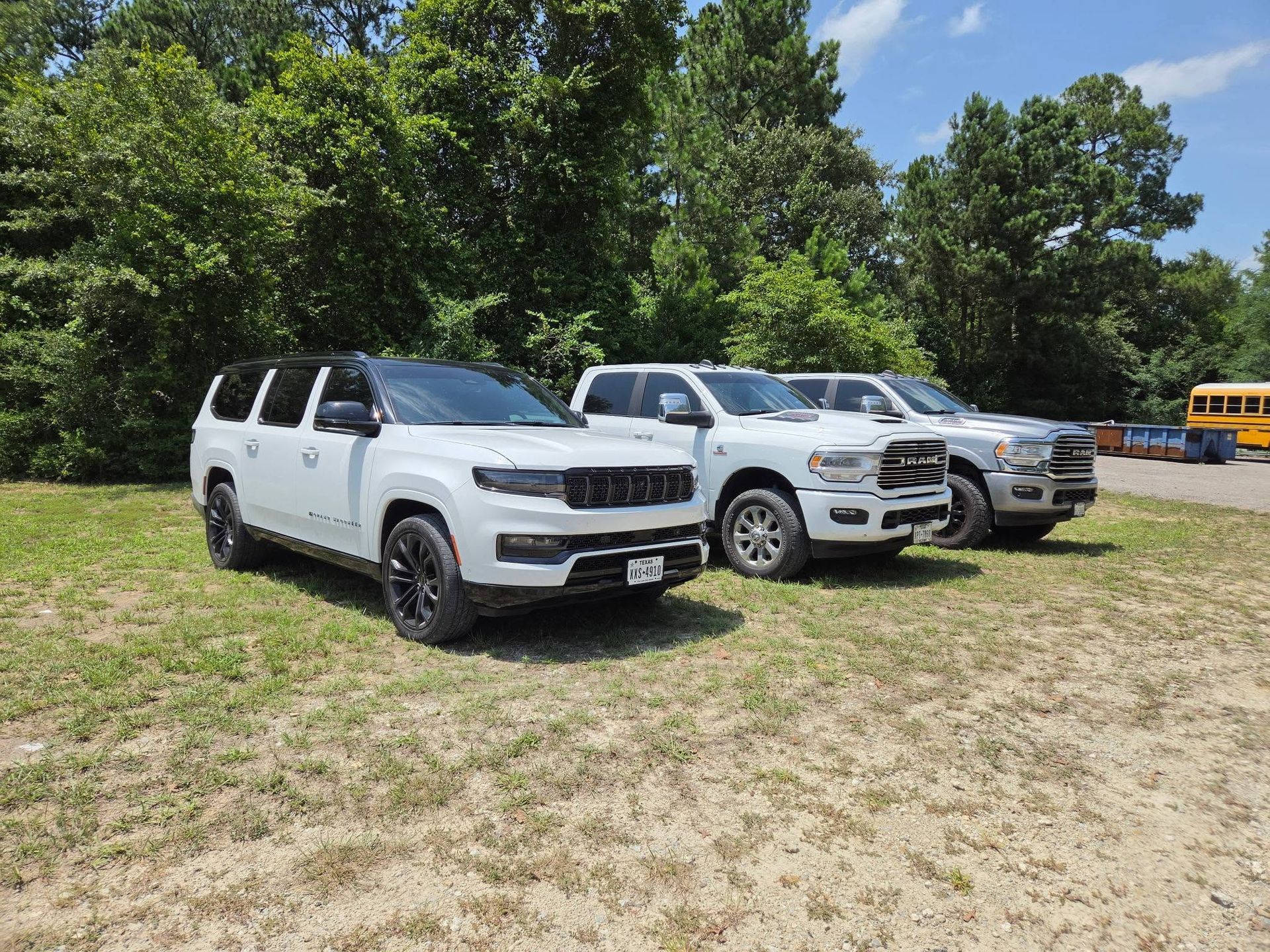 Three white SUVs parked on grass near trees, with a yellow school bus in the background