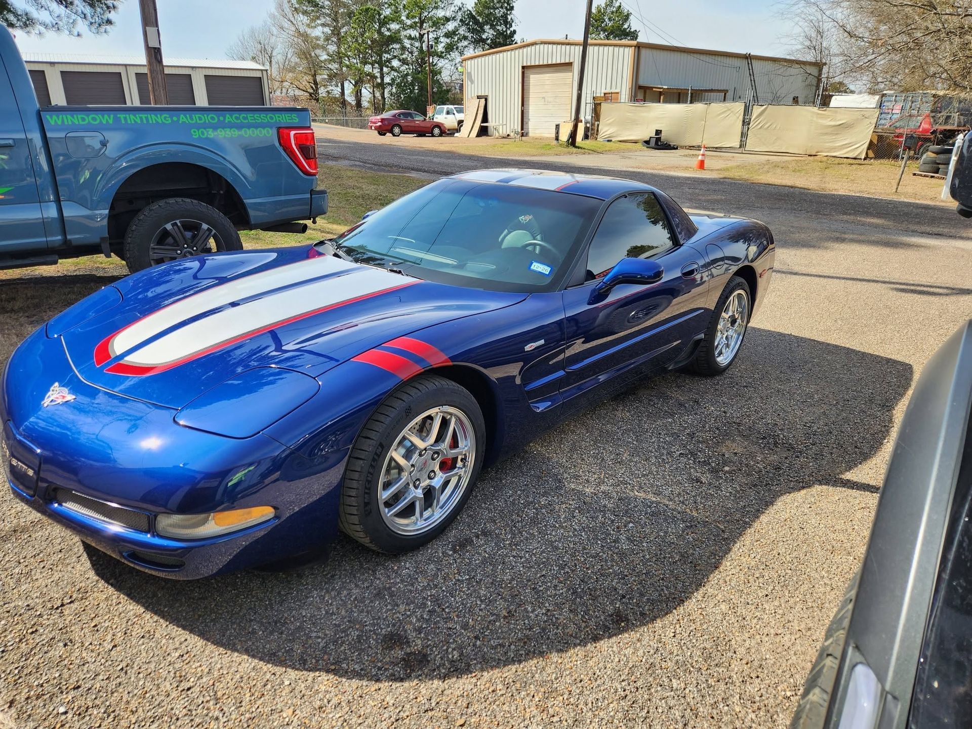 Blue sports car with red and white racing stripes parked on gravel near a building