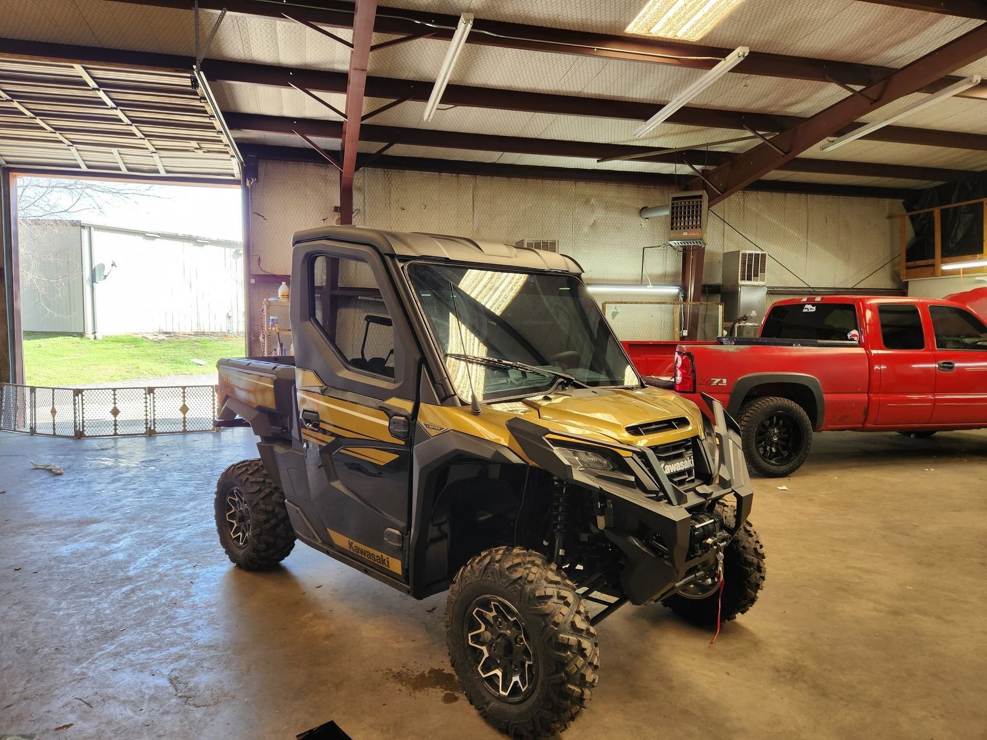 Damaged black-and-yellow off-road side-by-side in a garage with a red pickup truck nearby