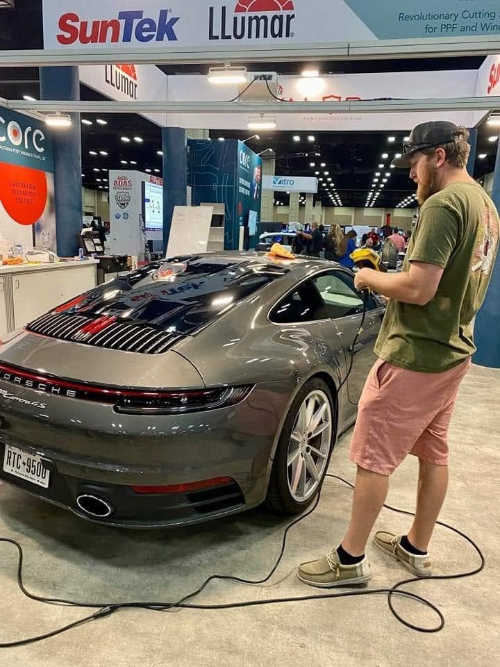 Man cleaning a gray sports car at an auto show booth under bright lights